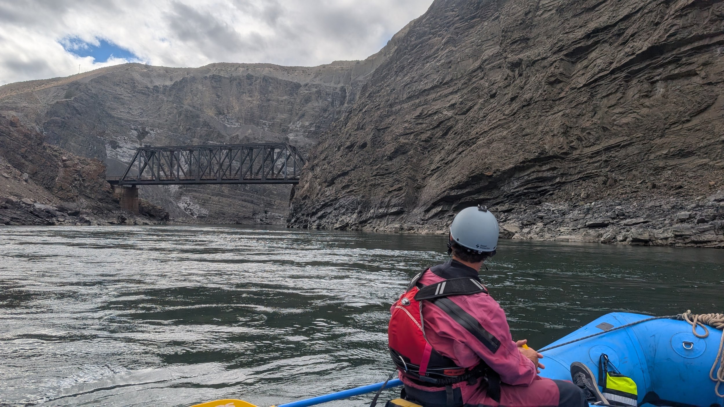 Person wearing a helmet and pink jacket sitting in a kayak on a river, with rocky cliffs and a metal bridge in the background.