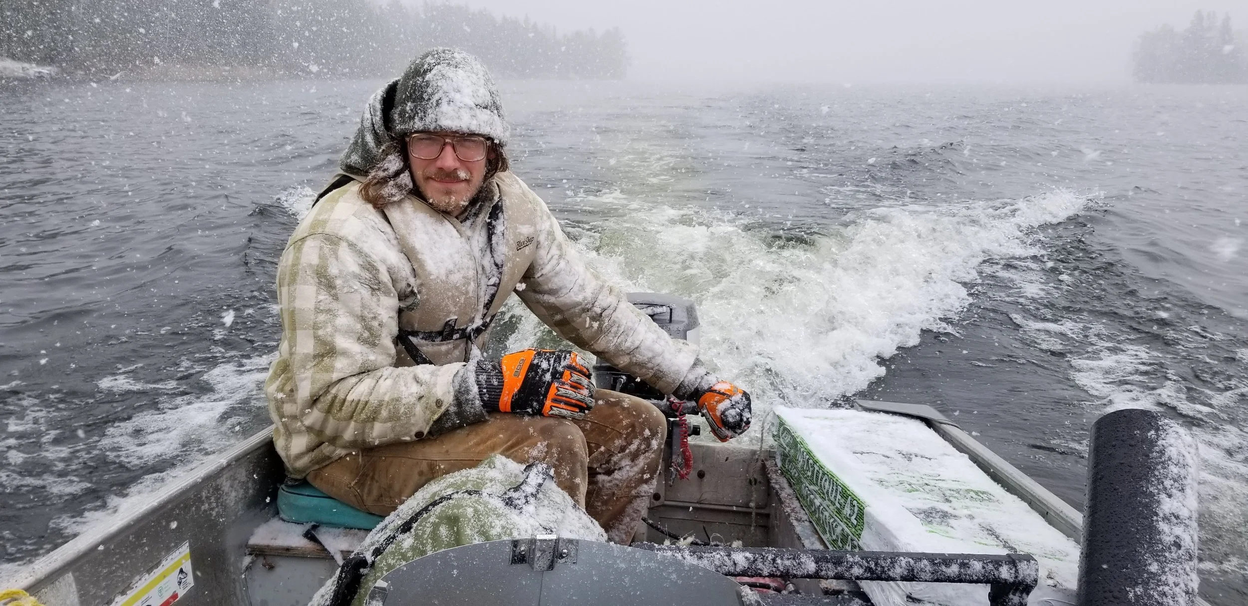 A person wearing a camouflage jacket, brown pants, orange gloves, and a snow-covered hat sitting in a small boat on a snow-covered body of water during a snowstorm, holding onto an orange safety lanyard attached to the motor.