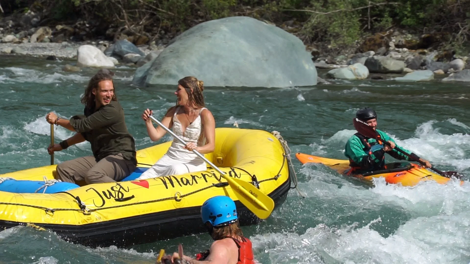 People enjoying white water rafting on a river, with two individuals sitting in a yellow raft and a person paddling an orange kayak nearby, surrounded by rocks and trees.
