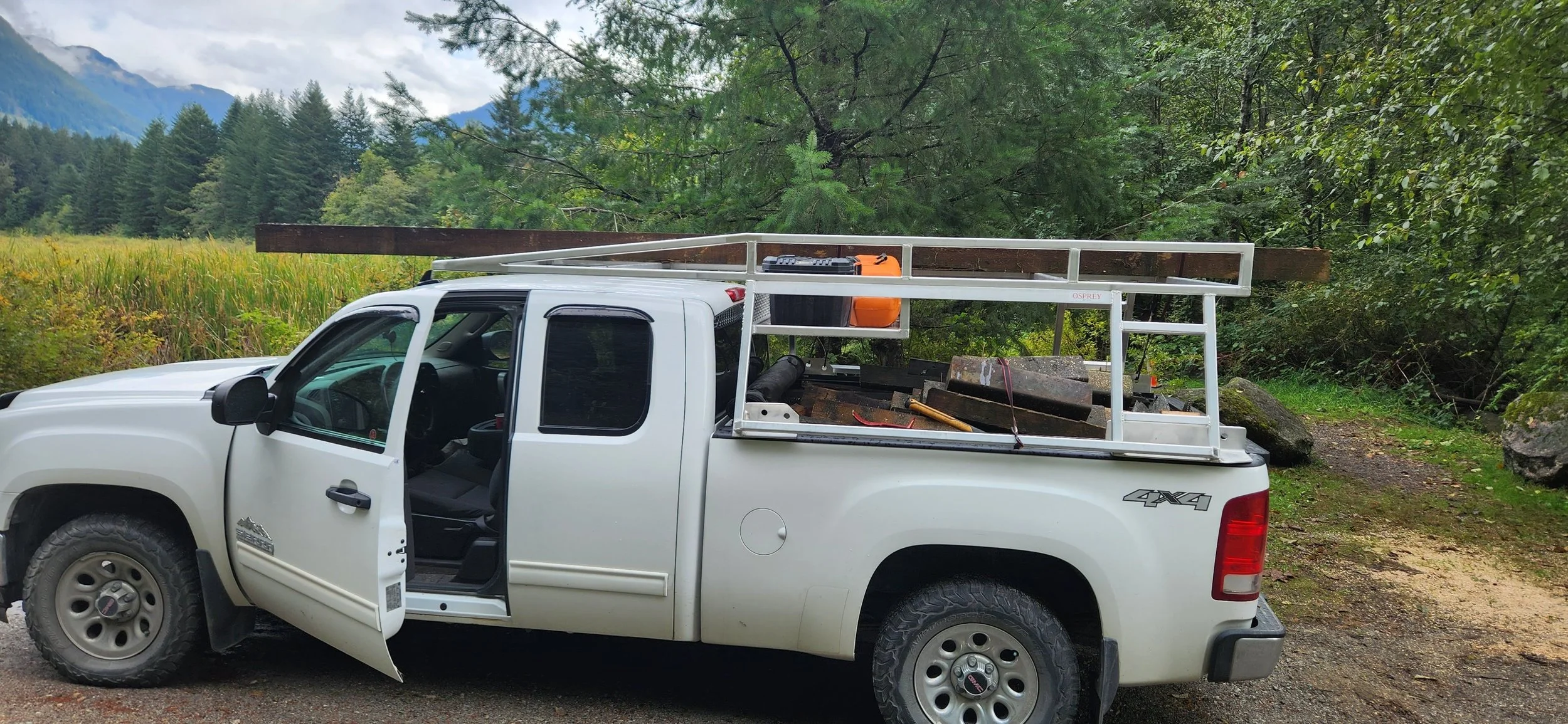 White pickup truck loaded with wooden beams and tools parked on a dirt path in a forested area with mountains in the background.