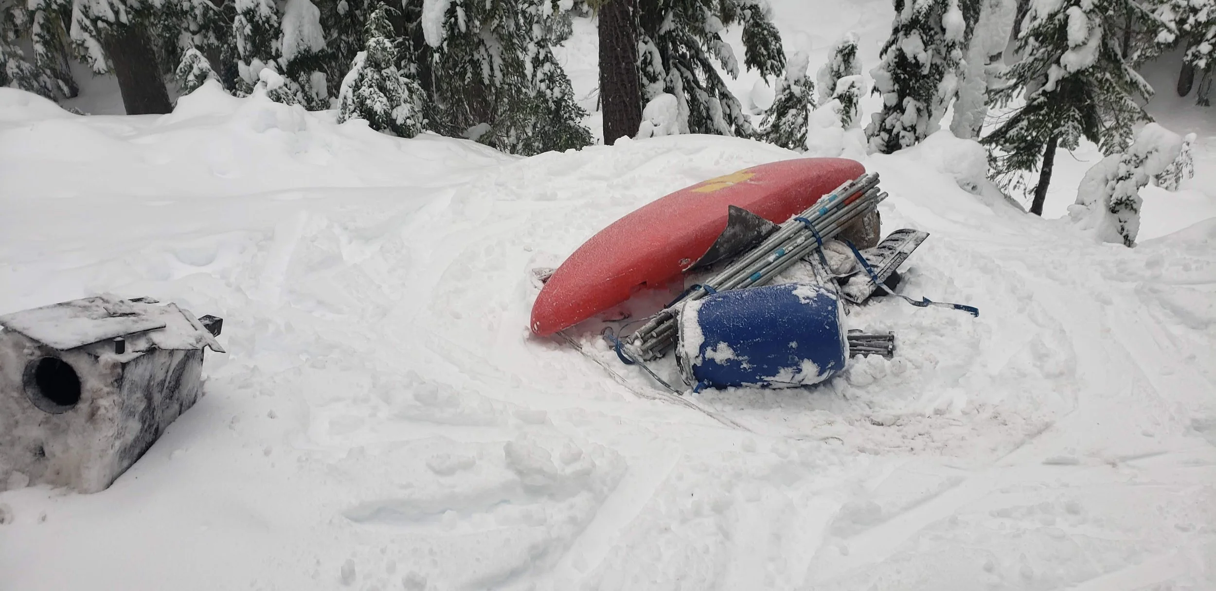 A snow-covered outdoor scene with a red sled, a pair of skis with poles, and a small wooden doghouse among snow-covered trees.