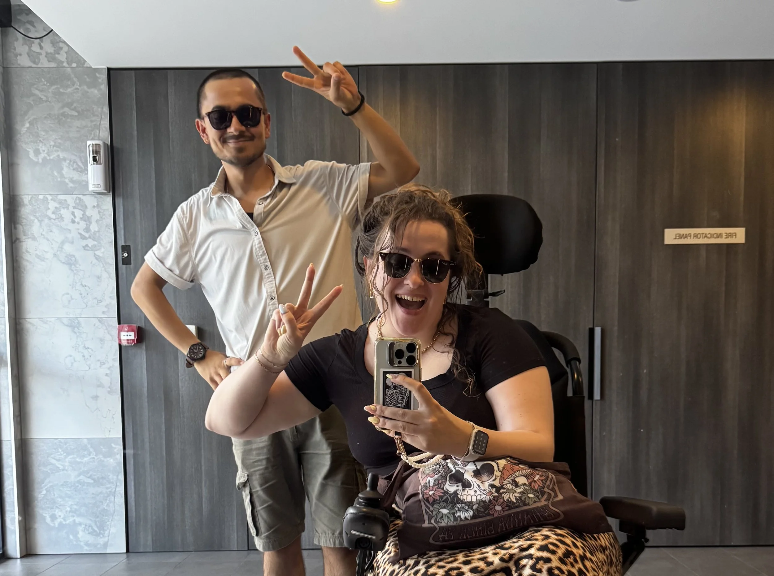Two people in an elevator taking a mirror selfie. The person in front is a woman with curly hair, wearing sunglasses, a black shirt, and a skincare tattoo on her chest, and she is making a peace sign with her hand. The person behind her is a man with short dark hair, wearing sunglasses and a white shirt, giving a peace sign with his right hand. The elevator has dark wood panels and a sign that says 'FIRE INDICATOR PANEL'.