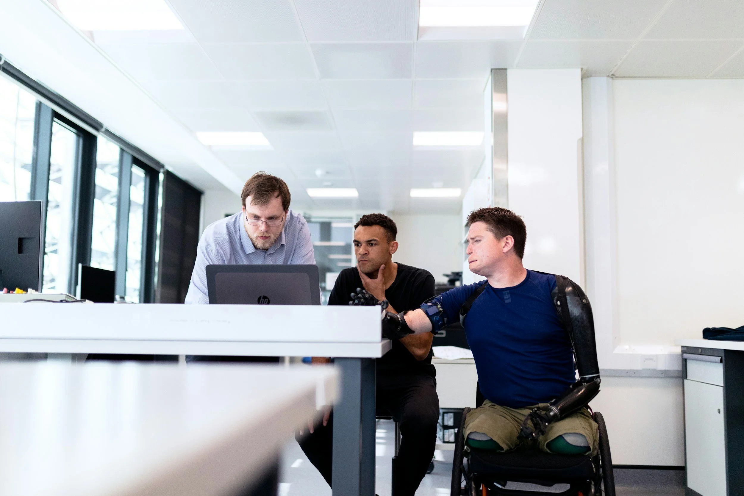 Three men in a modern office, two sitting at a white desk looking at a laptop, one standing leaning over to look at the same laptop. The office has large windows and white walls.