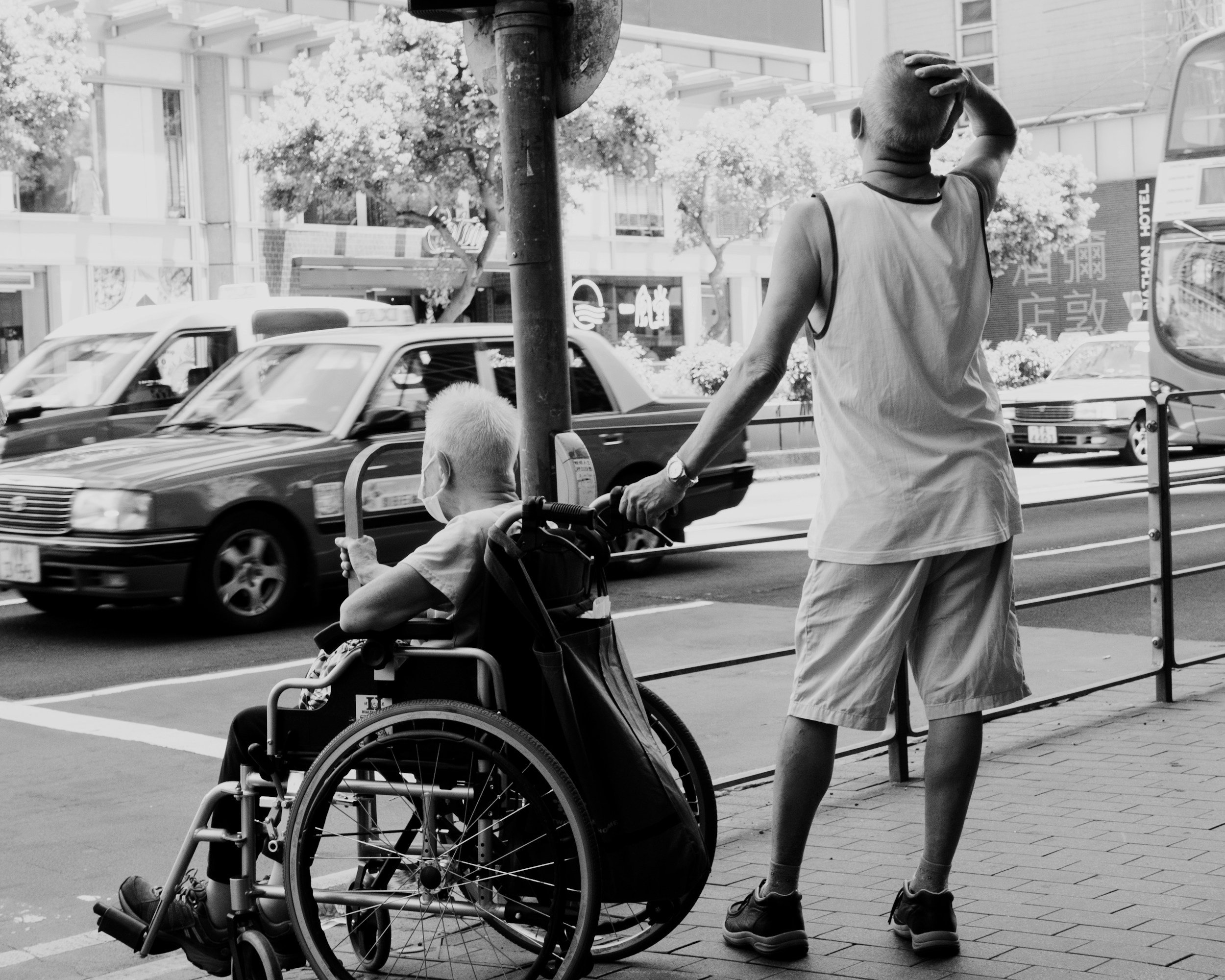 A young man walking on sidewalk, hand on head, next to a person in a wheelchair and a man pushing the wheelchair, all on a city street with taxi cabs and trees.