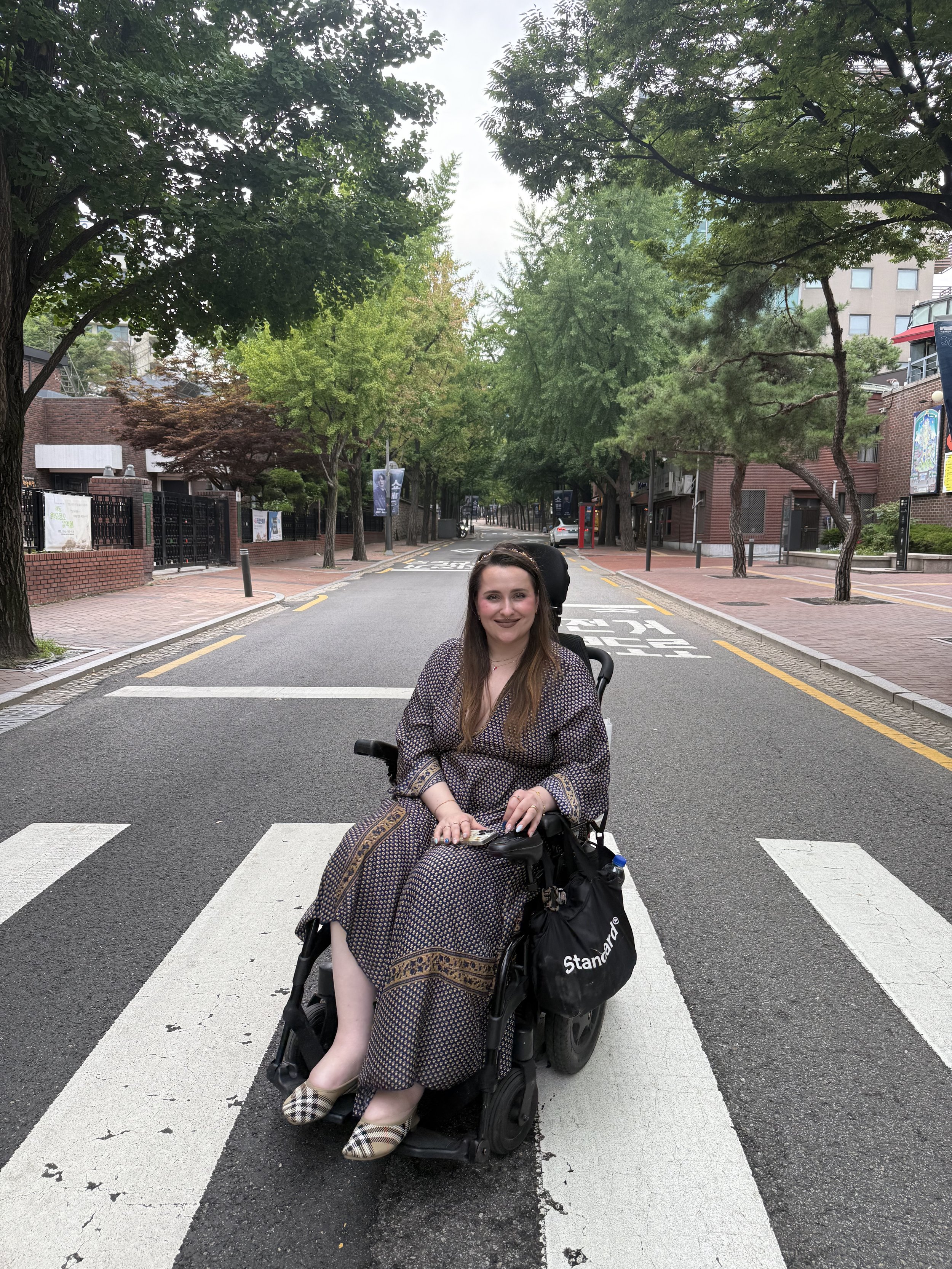 Woman sitting in a motorized wheelchair on a crosswalk in a city street, surrounded by trees and buildings.