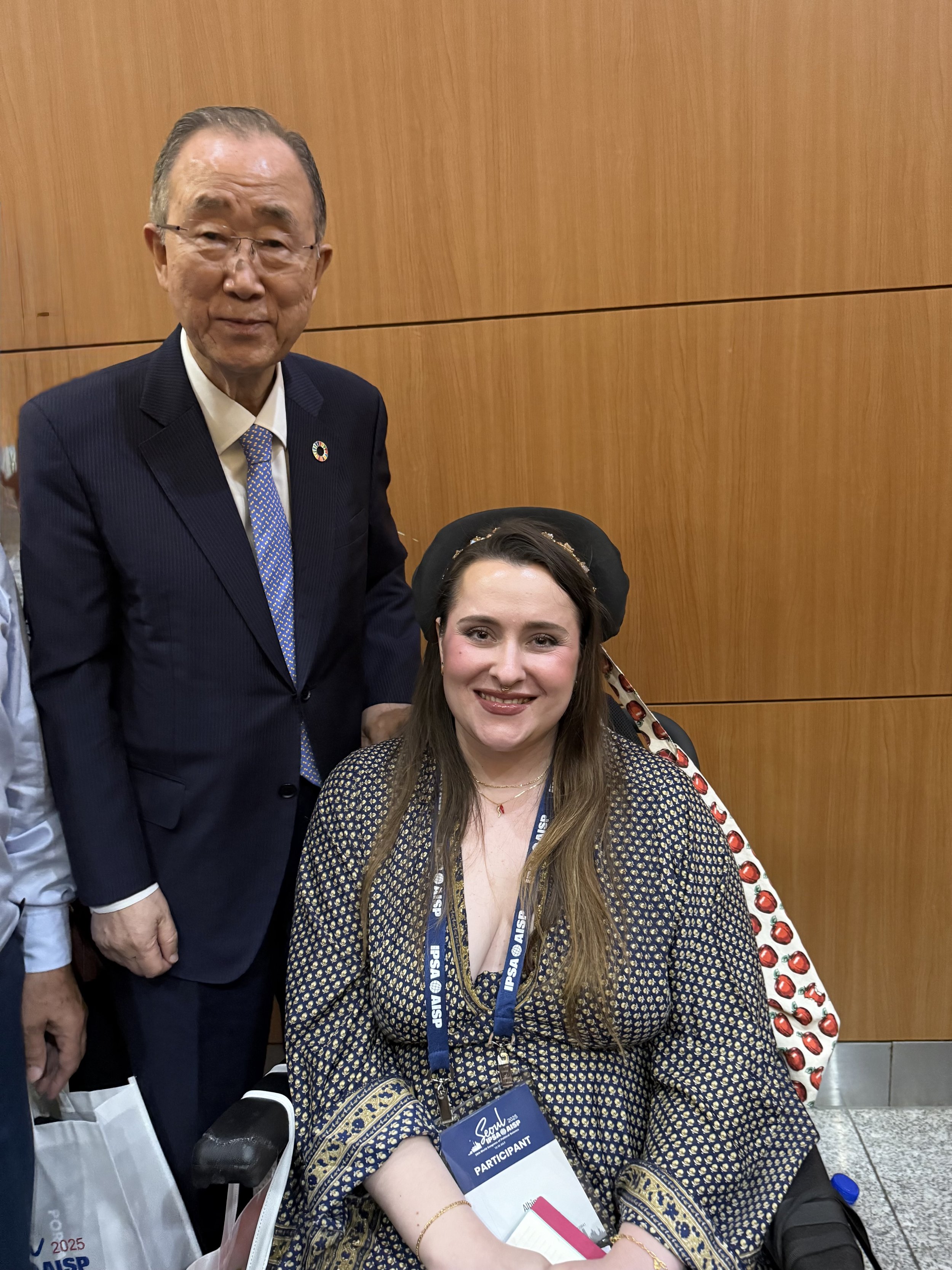An elderly man in a dark suit and glasses stands next to a young woman sitting in a wheelchair. The woman is smiling, has long brown hair, and wears a patterned dress with a name badge that reads "Participant." Both are at a conference or event with a wooden wall in the background.