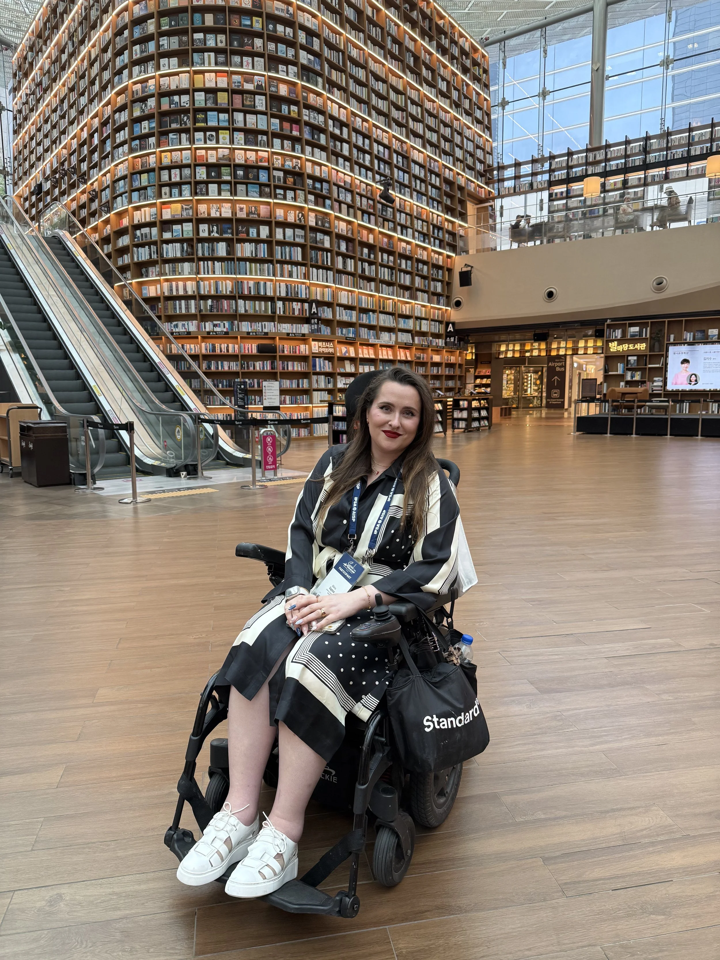 A woman sitting in a motorized wheelchair inside a modern bookstore or library with high ceilings, large glass windows, and tall bookshelves. She is wearing a black and white striped dress, white sneakers, and has long brown hair. She is smiling and holding her hands together, with various books and a water bottle attached to her wheelchair. In the background, there are escalators, people sitting at a balcony, and a digital display screen.
