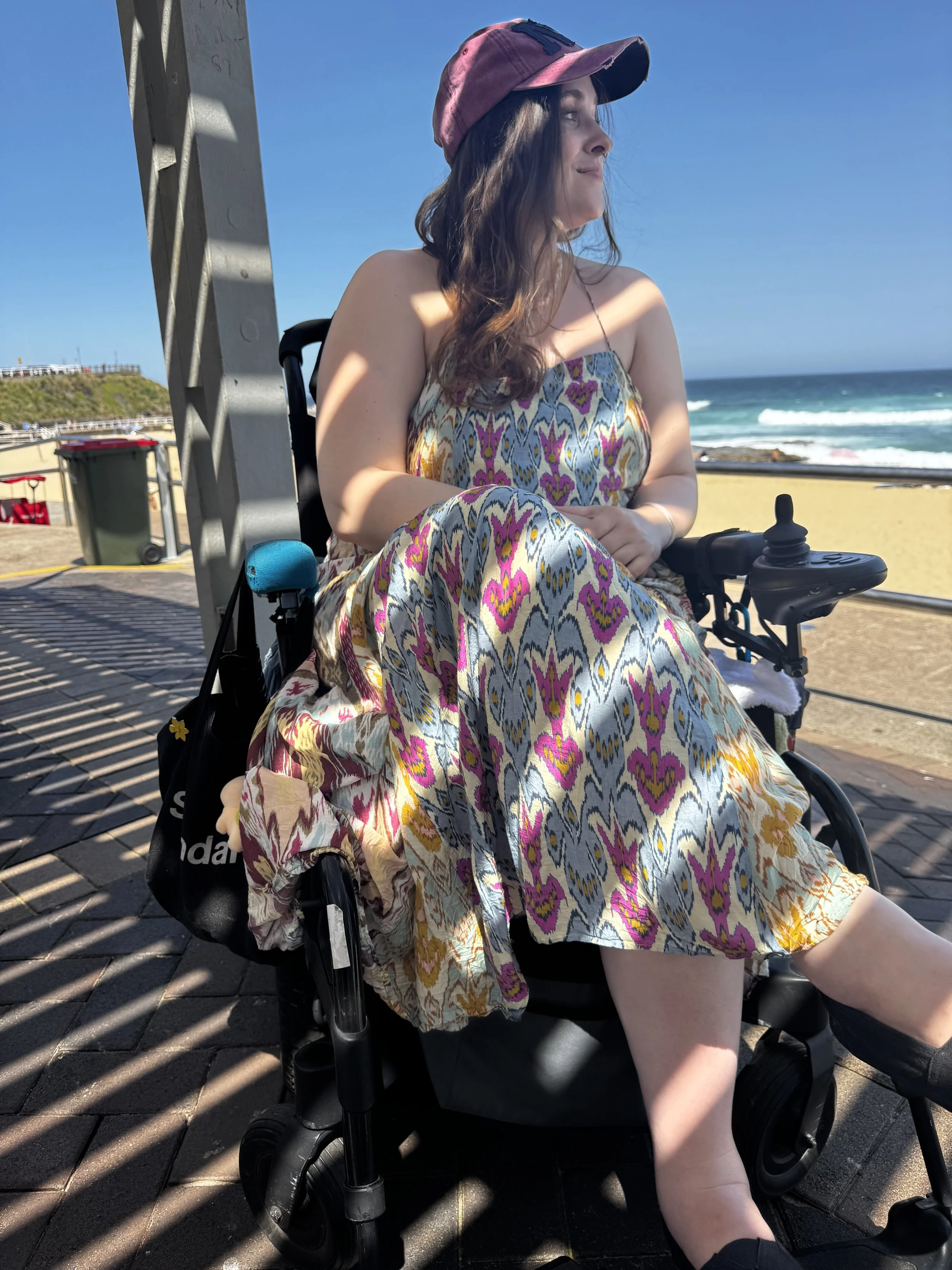 Woman with dark hair wearing a pink cap and a patterned dress sitting in a wheelchair at the beach, overlooking the ocean with rocky shoreline and blue sky in the background.