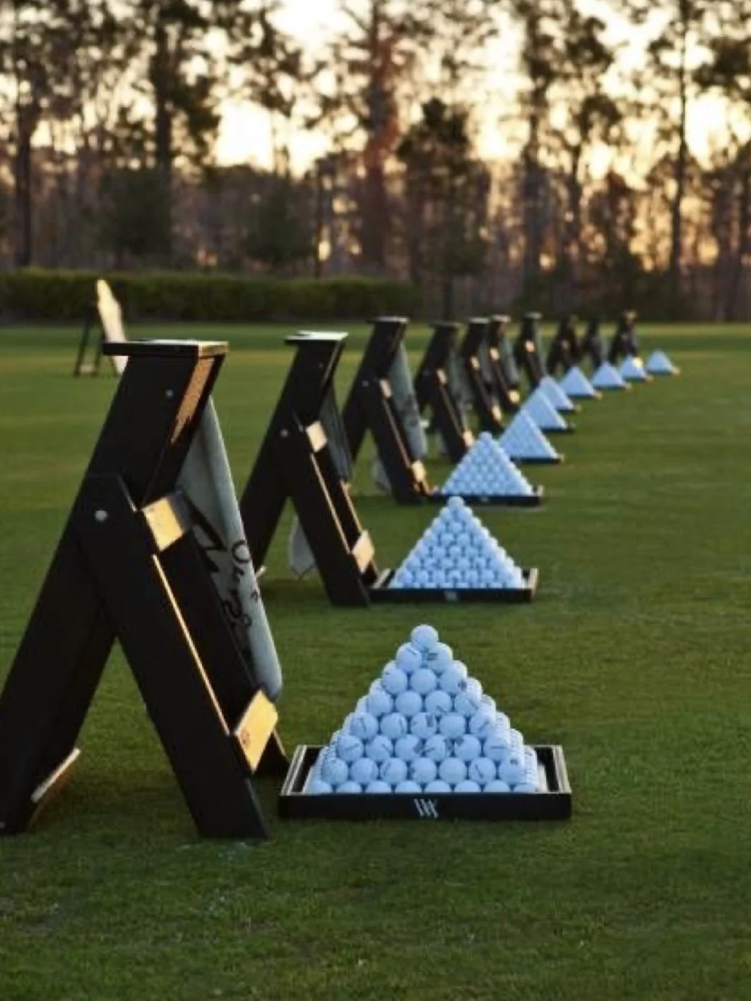 Golf driving range with wooden stands and neatly arranged white golf balls in pyramid shapes, set on a well-maintained grassy field during sunset.