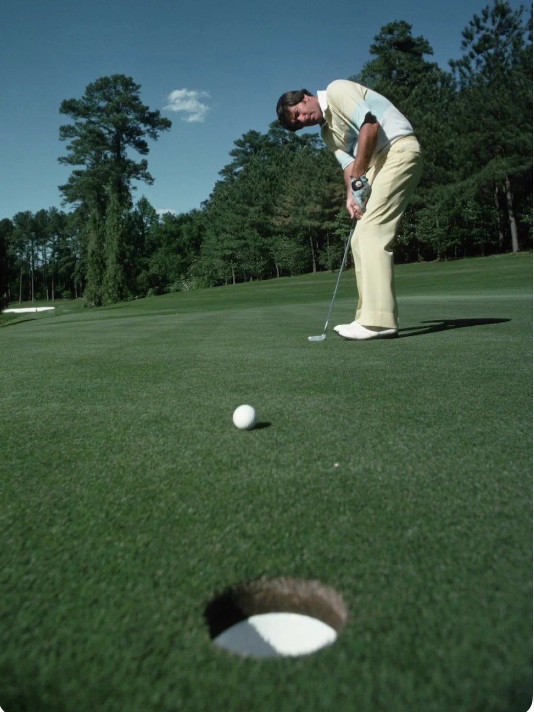 A man in light-colored clothing prepares to putt a golf ball on a golf course, with the hole visible in the foreground and trees in the background under a clear blue sky.