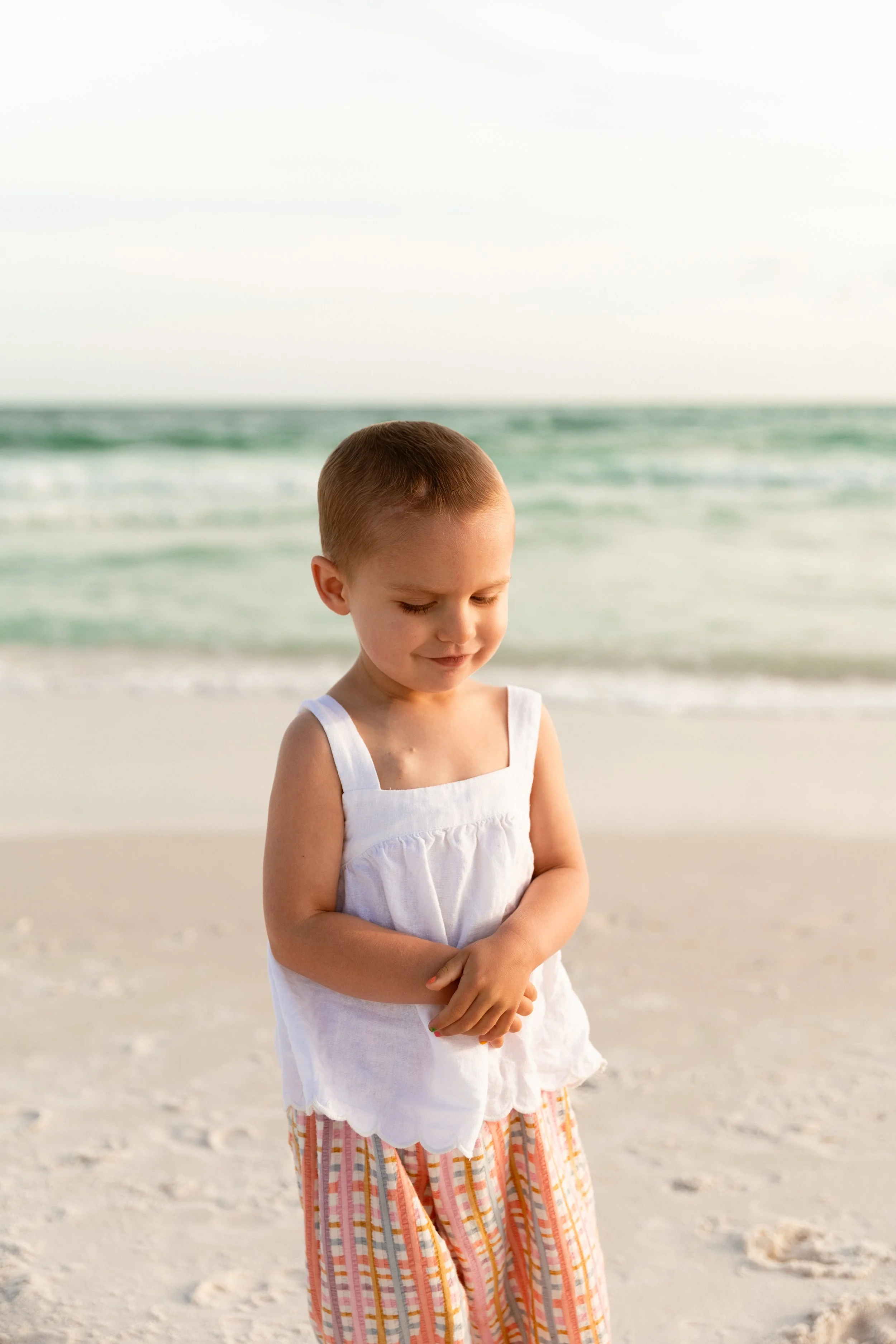 A young girl with short hair and a white dress standing on the beach with the ocean in the background.