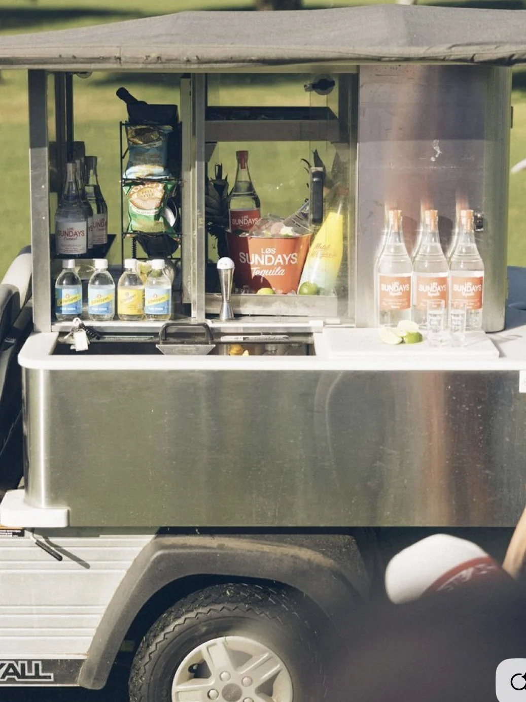 A small mobile bar cart with a silver metal exterior, stocked with bottled water, lime wedges, liquor bottles labeled 'Sundays,' and other bar supplies, set on a grassy outdoor area.