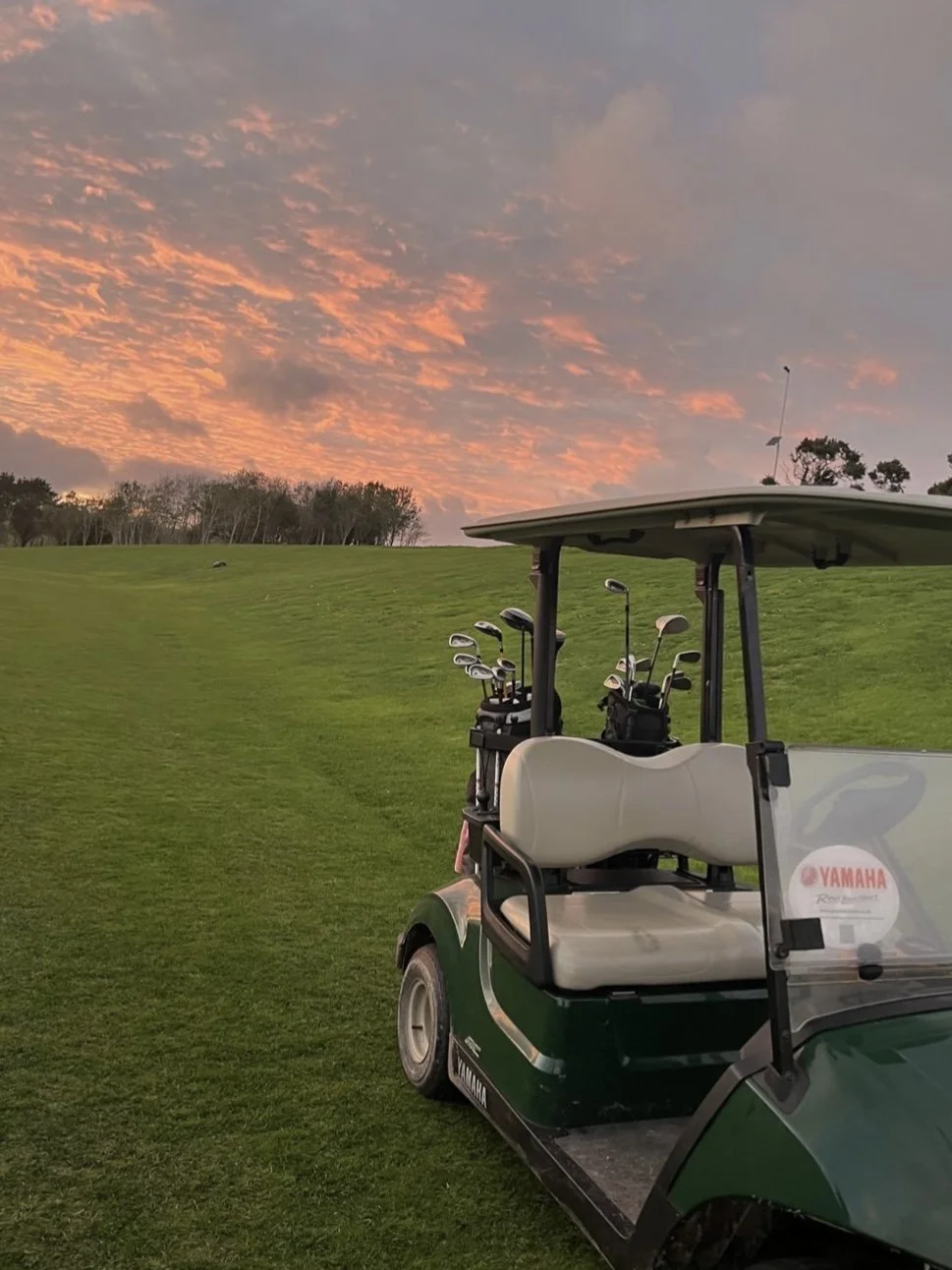 A green golf cart with multiple golf clubs in the holder, parked on a well-manicured golf course with a sunset sky.
