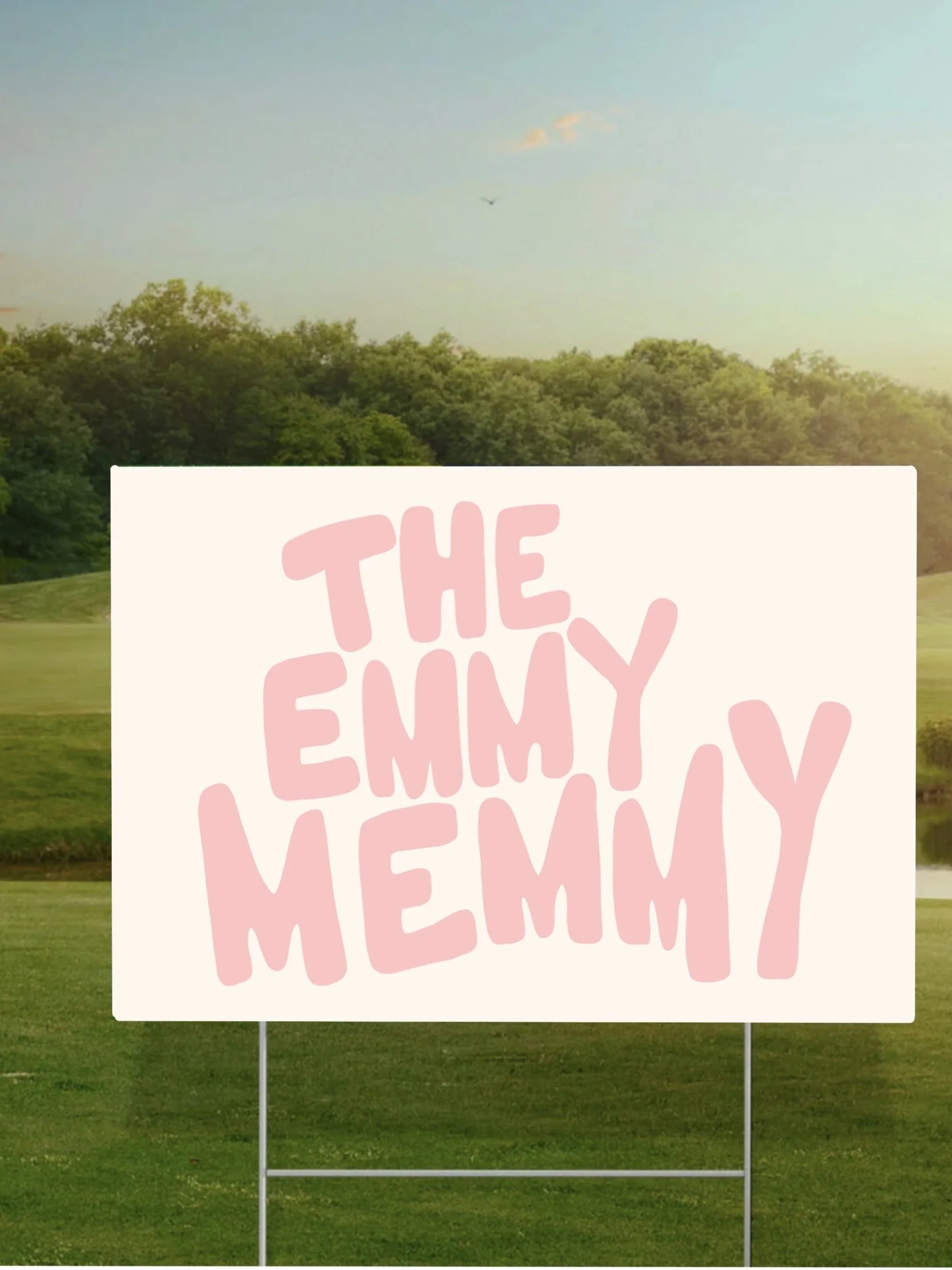 A sign with pink text reading 'The Emmy Memmy' is placed on a grassy field with trees in the background and a blue sky overhead.