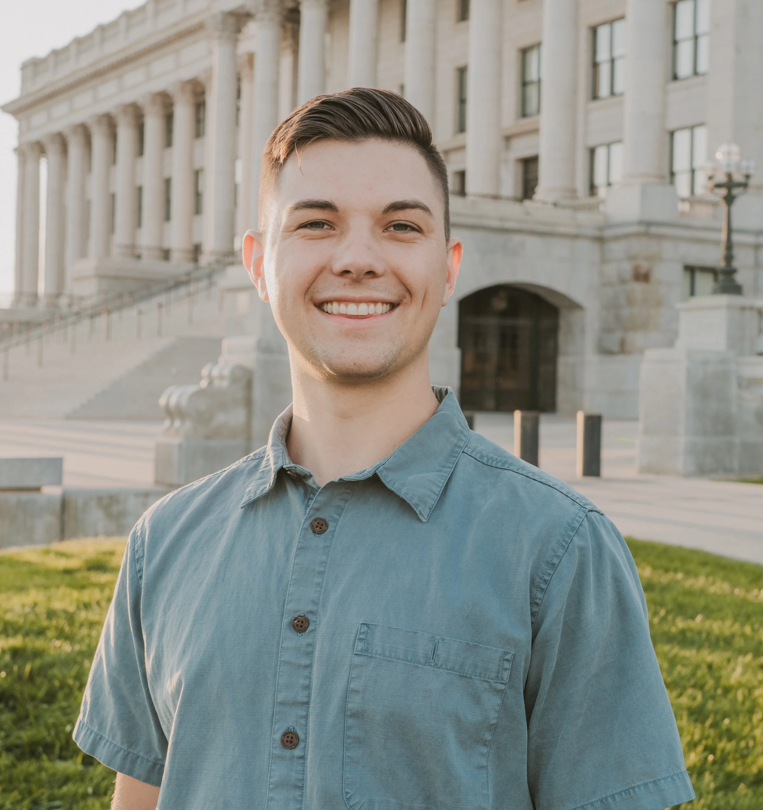 Close-up of a smiling young man with slicked-back hair wearing a blue button-up shirt outdoors with a large historical building in the background.