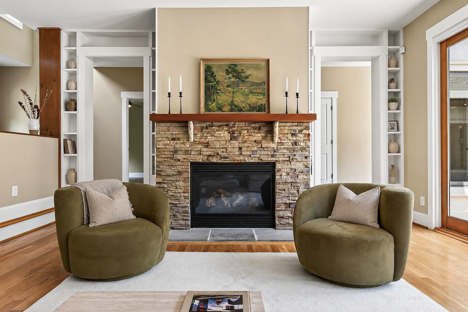 Living room with a fireplace, two olive green armchairs with beige pillows, wooden flooring, beige walls, and built-in white shelves on either side of the fireplace. A painting hangs above the fireplace, and a sliding glass door is on the right.