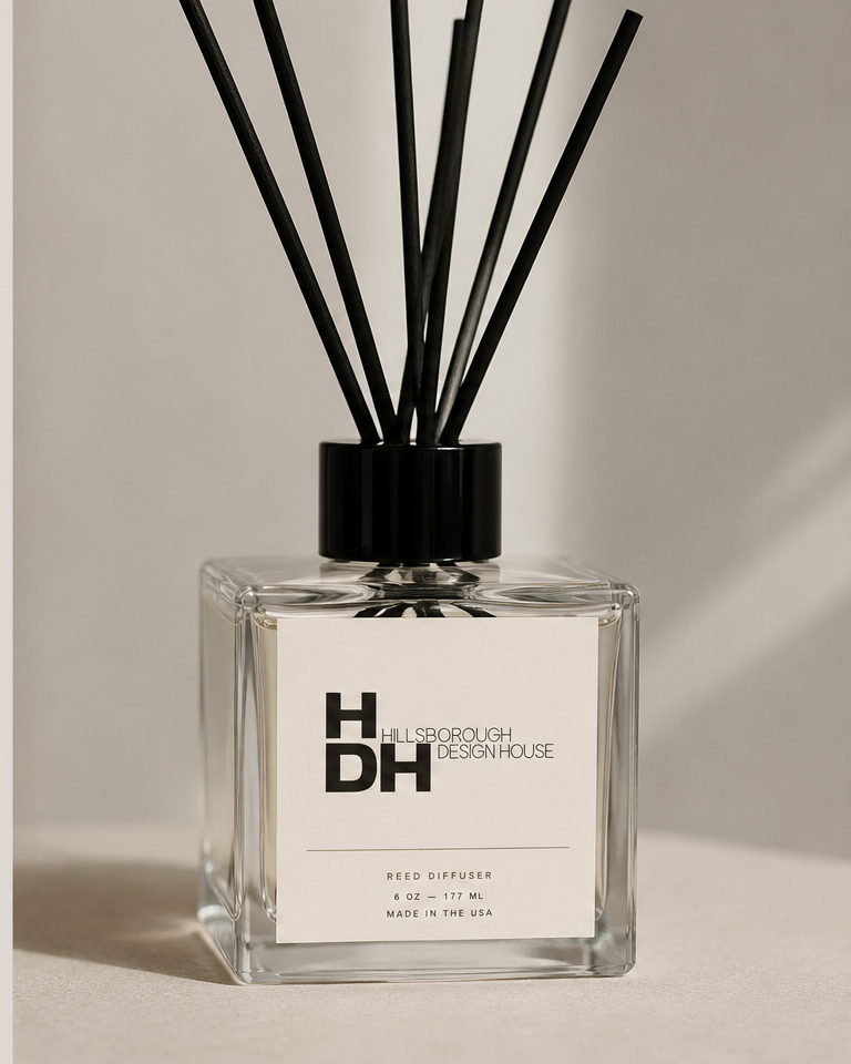 A clear glass reed diffuser with black reeds and a white label showing 'Hillsborough Design House,' 'Reed Diffuser,' and details about size and origin.