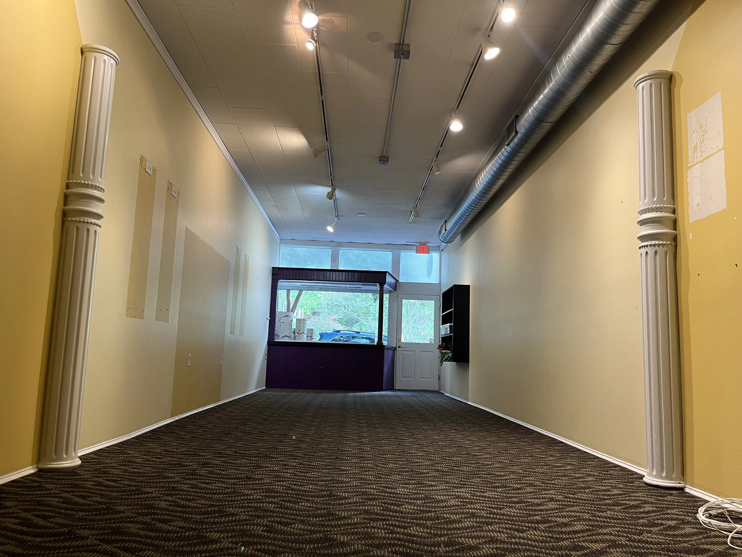 Interior of an empty commercial space with yellow walls, decorative pillars on the sides, track lighting on the ceiling, and a large window at the back showing an outdoor view.
