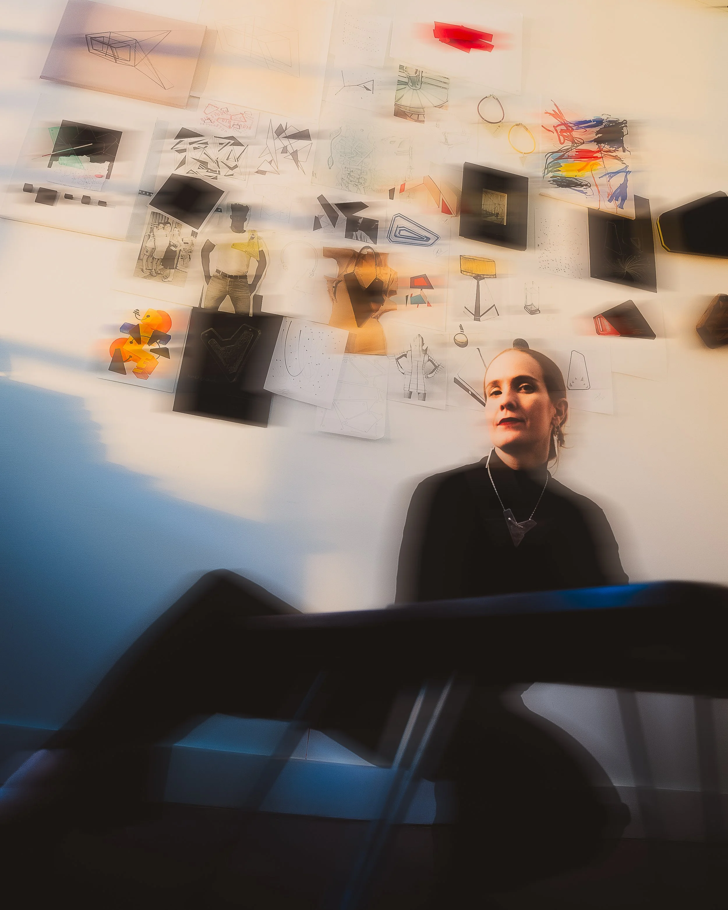 A person is taking a photo of a wall covered with sketches, drawings, and various paper notes related to design and architecture. The person is partially visible at the bottom right corner, wearing a black shirt and a necklace.