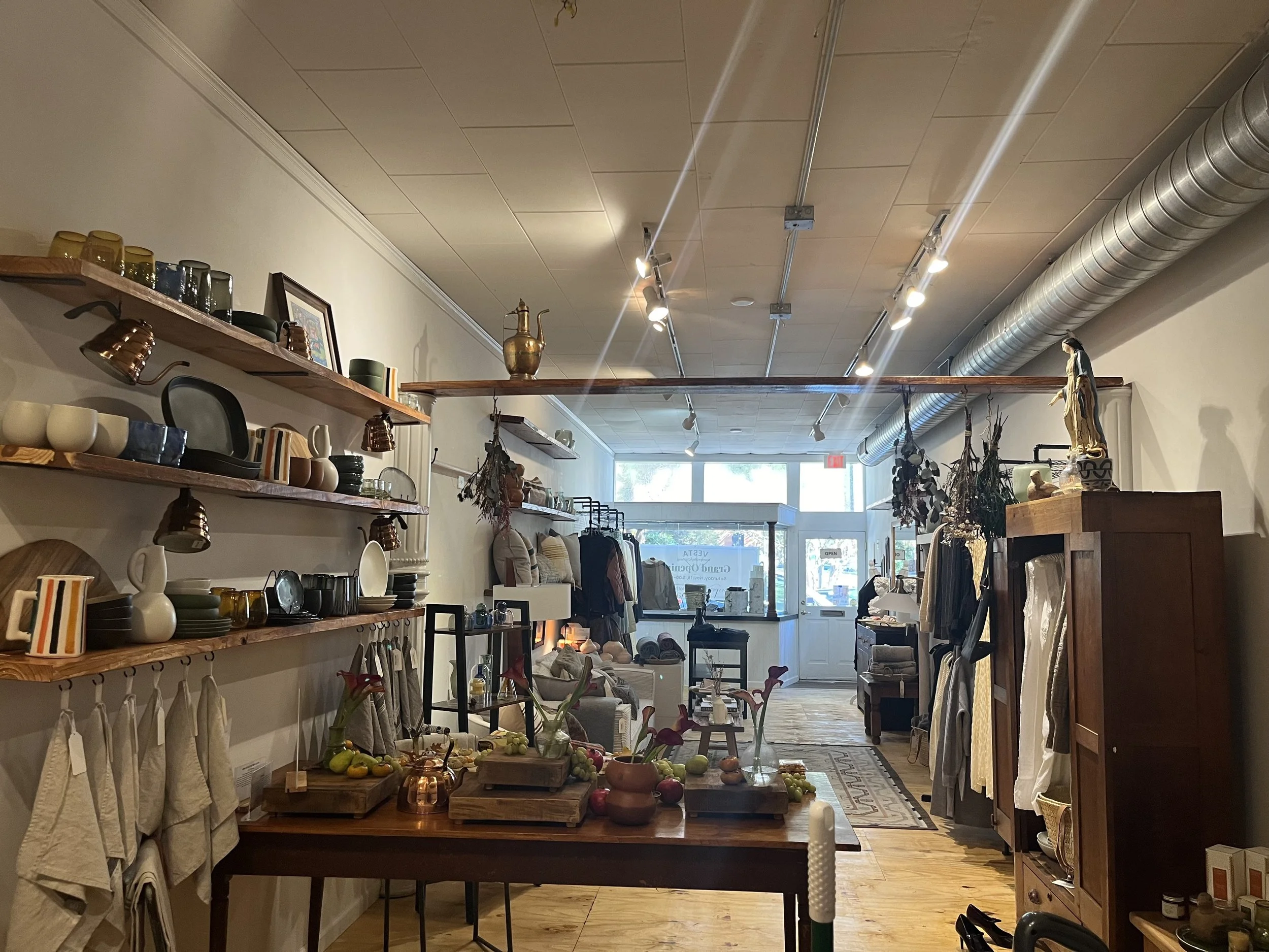 Interior view of a home decor and gift store. Wooden shelves display dishes, cups, and decorative items, with a table in the center holding fruits and plants. Racks and displays along the walls showcase various textiles and home accessories. Large windows at the back let in natural light.