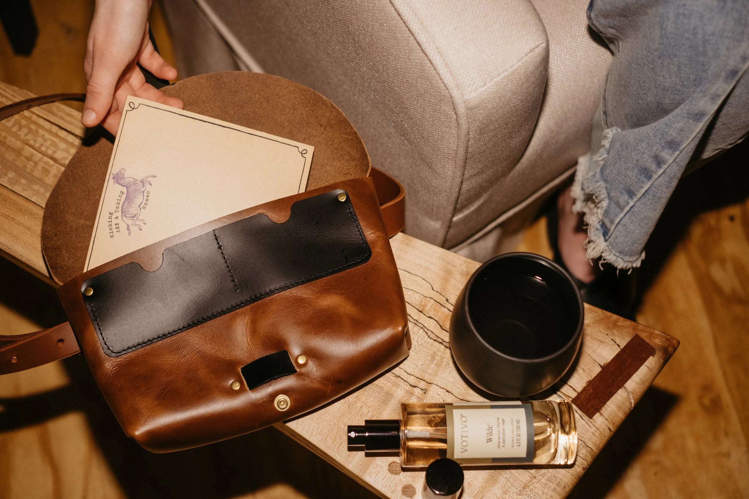 Close-up of a table with a brown leather bag, a perfume bottle labeled 'VOTIV' by 'Wilde', a black cup, and some papers, with a person's hand reaching for a paper in the background.