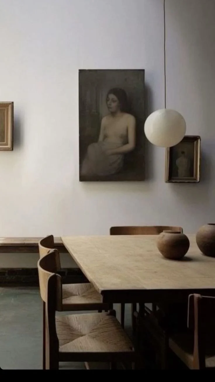A dining room with a wooden table, four chairs, and three pottery vases on the table. The wall has framed artworks and a round white paper lantern hanging near a large framed black-and-white photo of a topless woman sitting with her arms crossed.