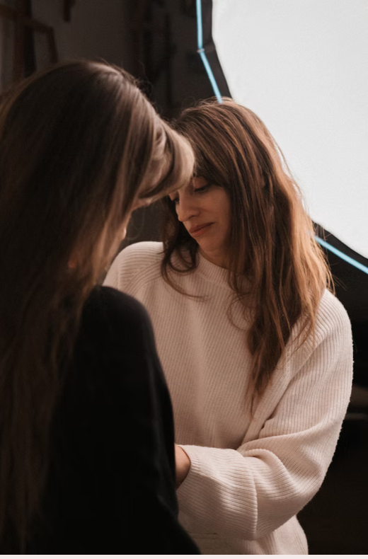 Two women with brown hair are close together, one wearing a white sweater and the other with long brown hair, in front of a photography studio light and backdrop.