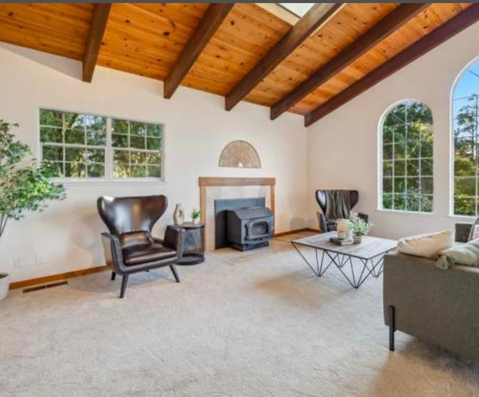 Living room with beige carpet, two dark leather armchairs, a grey couch, a coffee table, and a fireplace under a wooden vaulted ceiling with large windows providing natural light.