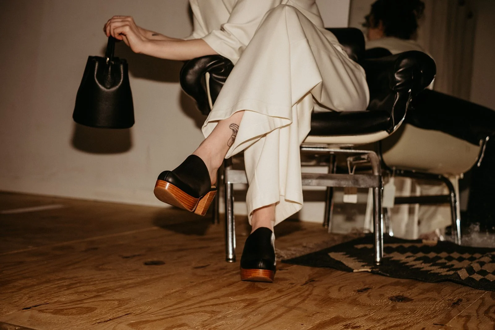 Person sitting on a black leather vintage bench, wearing beige pants and black platform shoes with wooden soles, holding a black handbag, with another person blurred in the background.