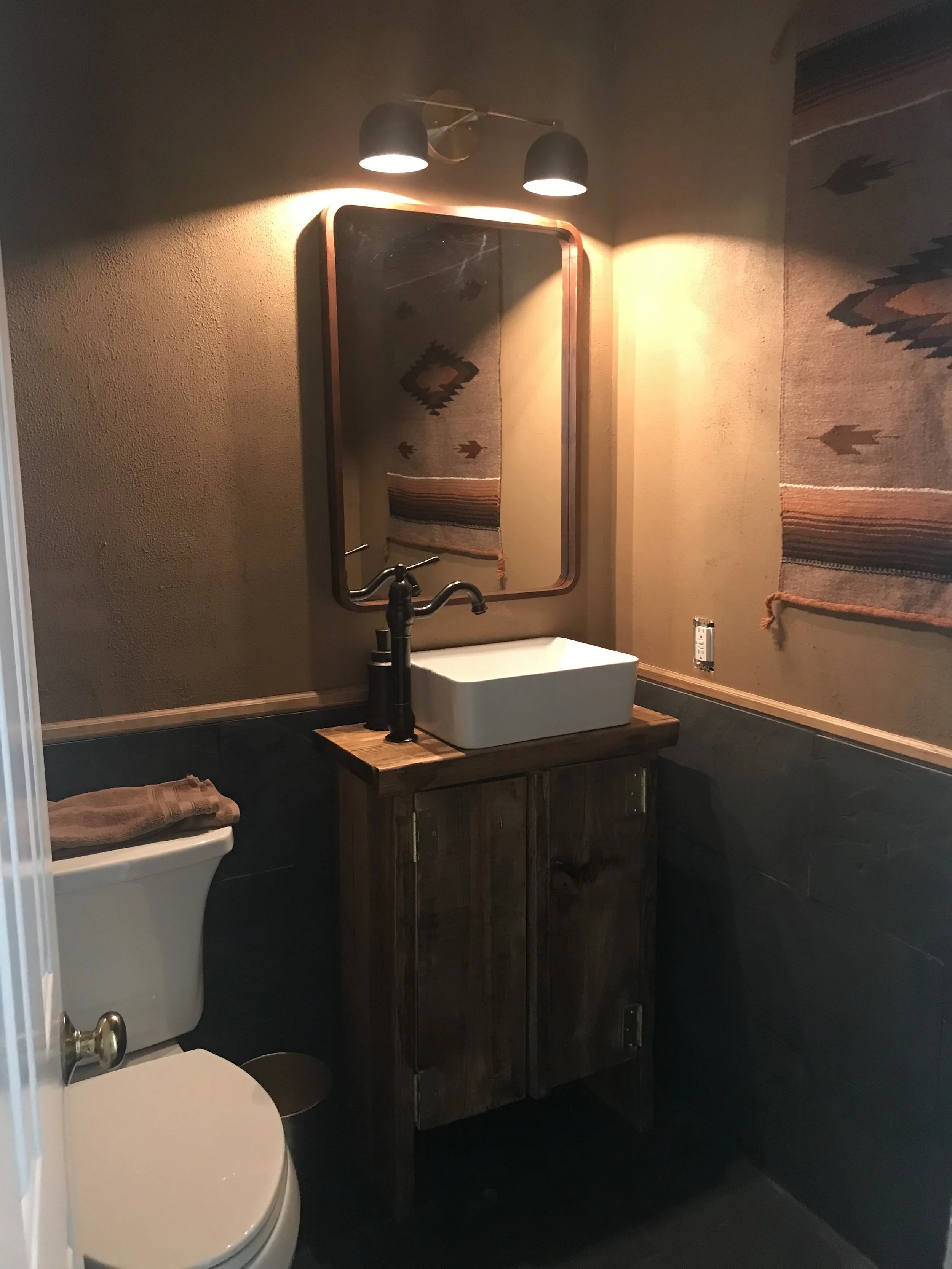A bathroom with a modern white rectangular sink on a wooden vanity, a mirror above the sink, a wall-mounted light fixture with two black shades, a beige wall hanging with southwestern patterns, a toilet with a brown towel, and dark tiling on the lower walls.