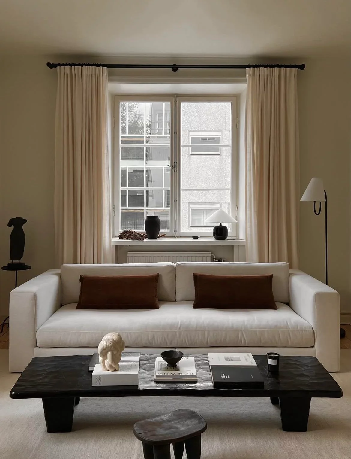 Living room with a white sofa, brown pillows, black coffee table with decorative objects, large window with beige curtains, and modern lamps.