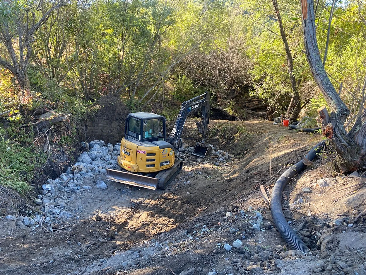 Compact excavator reshaping the creek channel beneath streamside trees