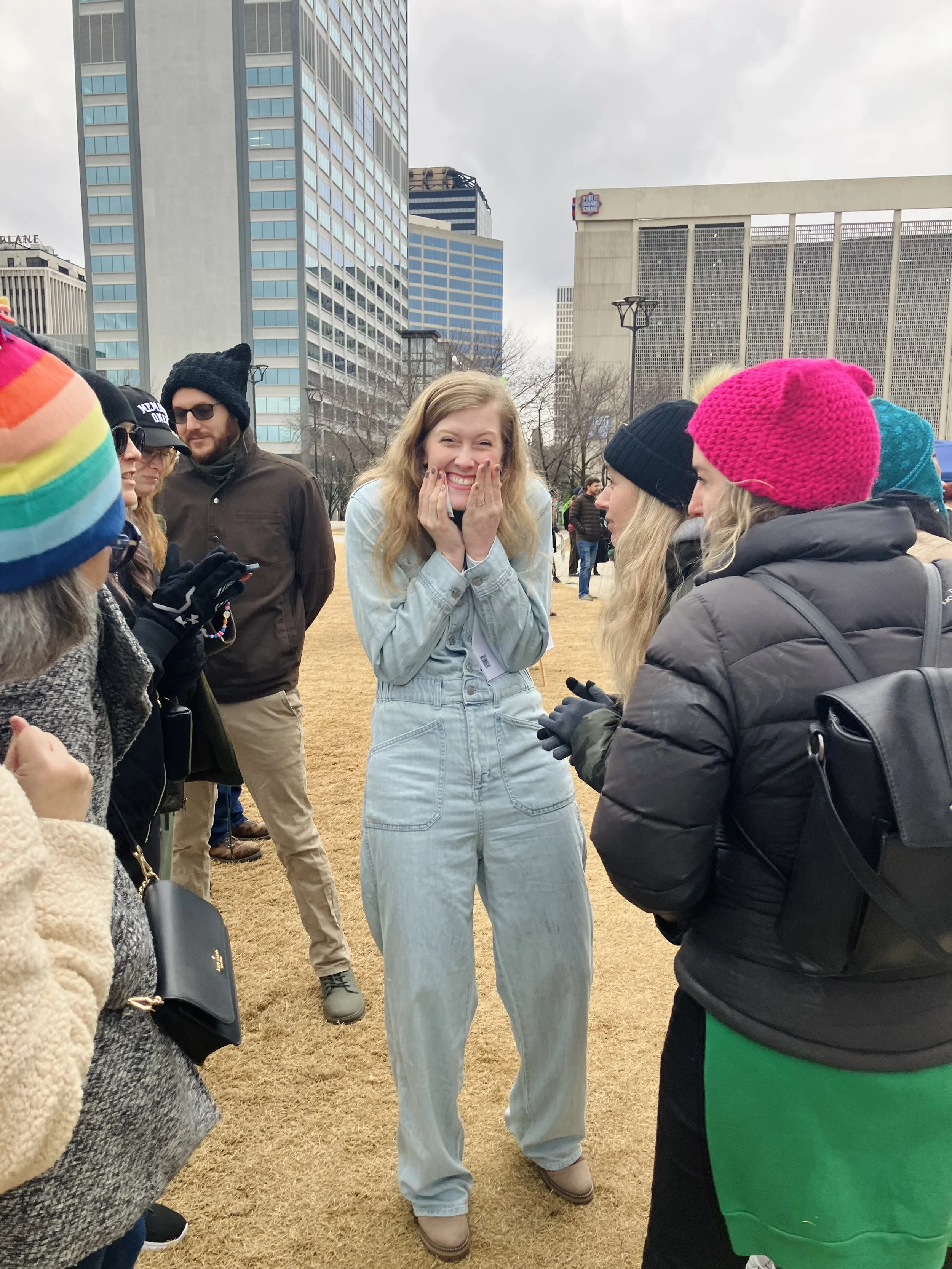 A group of people gathered outdoors on a cloudy day in an urban setting with tall buildings in the background, including a woman with curly blonde hair and a light denim jacket smiling and holding her face with both hands.