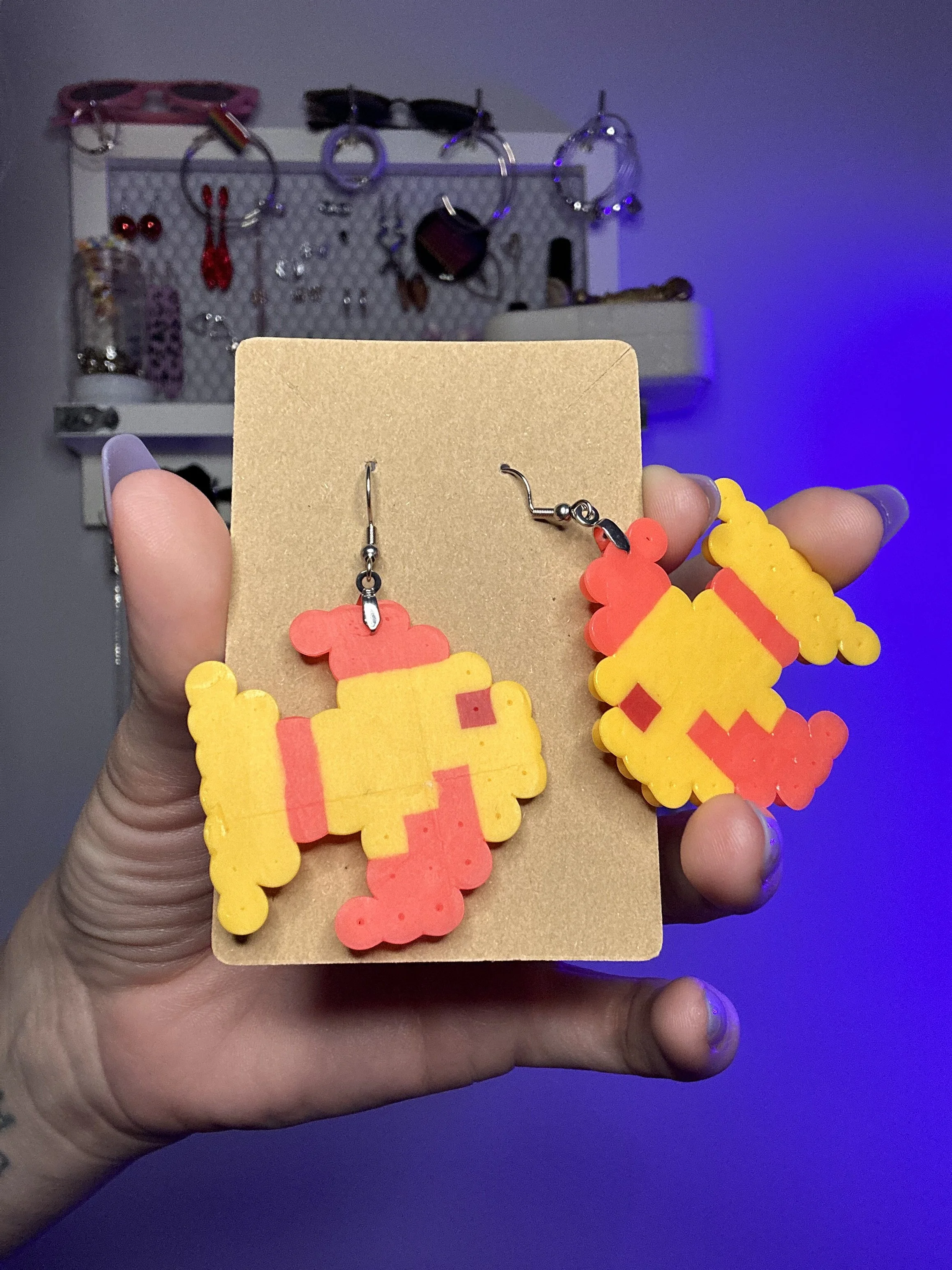 Hand holding a pair of yellow and orange fish earrings, with blurred jewelry organizer in the background.