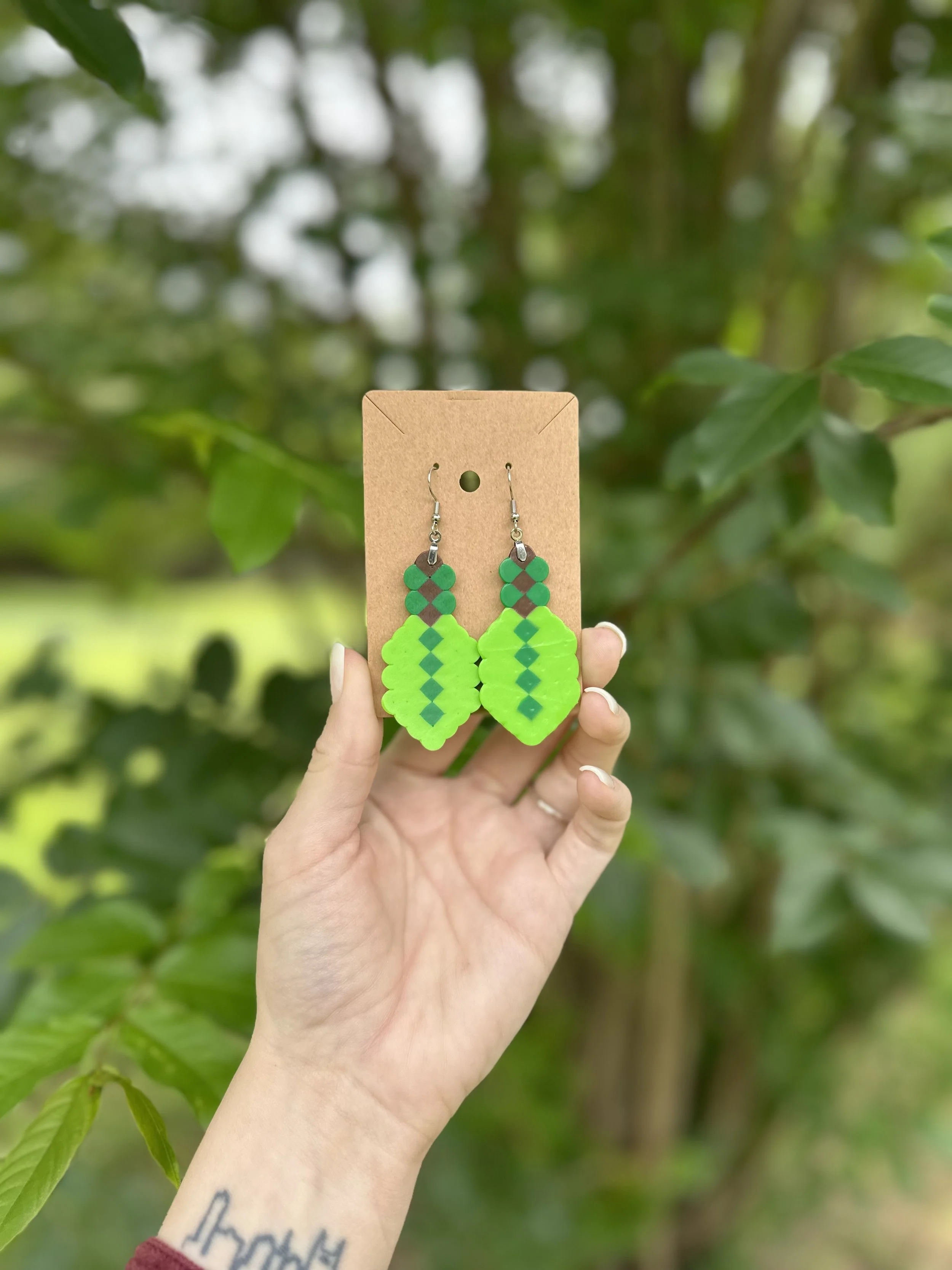 Hand holding a pair of green beaded earrings against a blurred green leafy background.