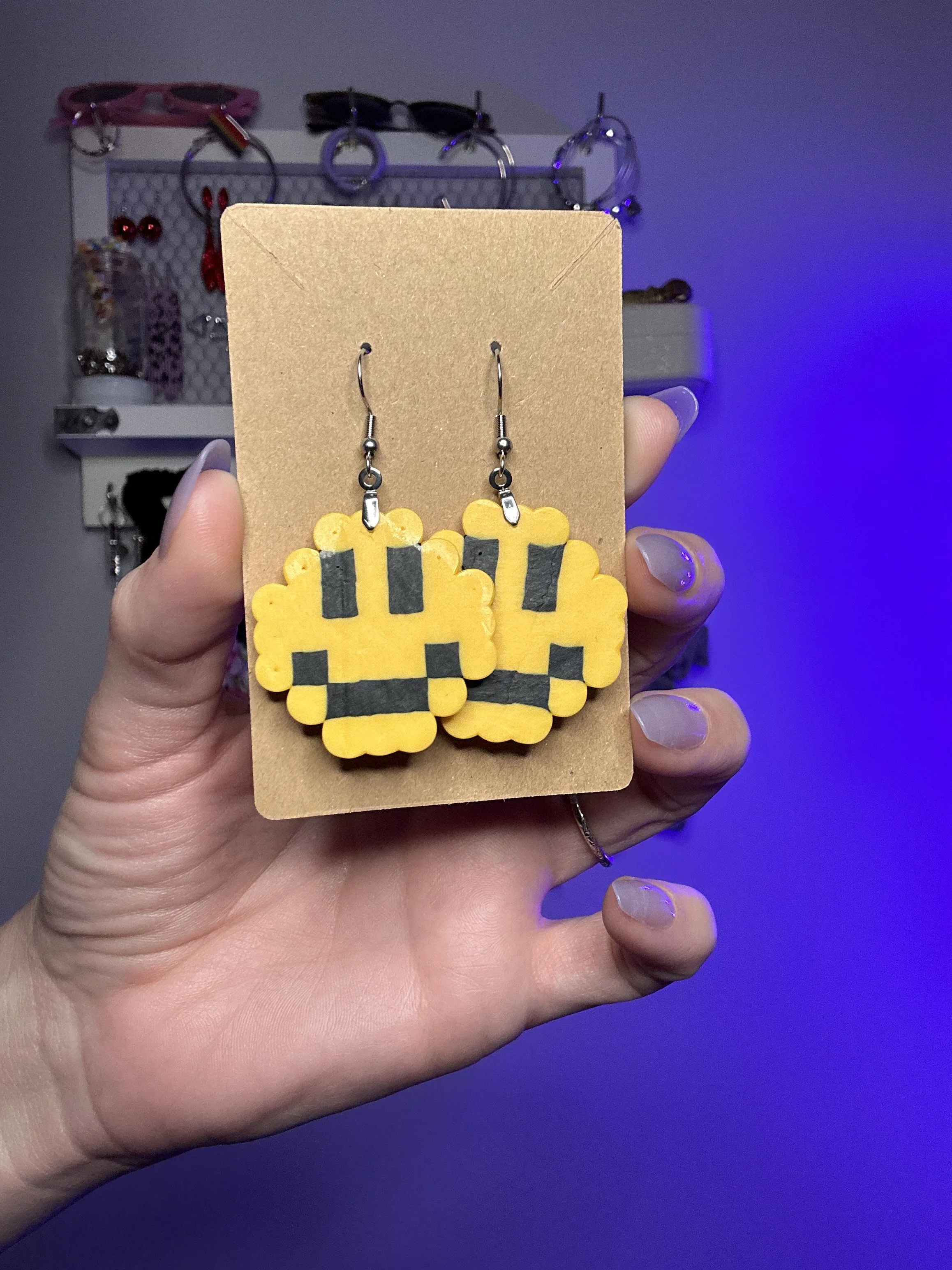 A hand holding a cardboard display with yellow and black earrings shaped like pixelated cacti. The background includes a hook rack with various accessories, with purple lighting affecting the scene.