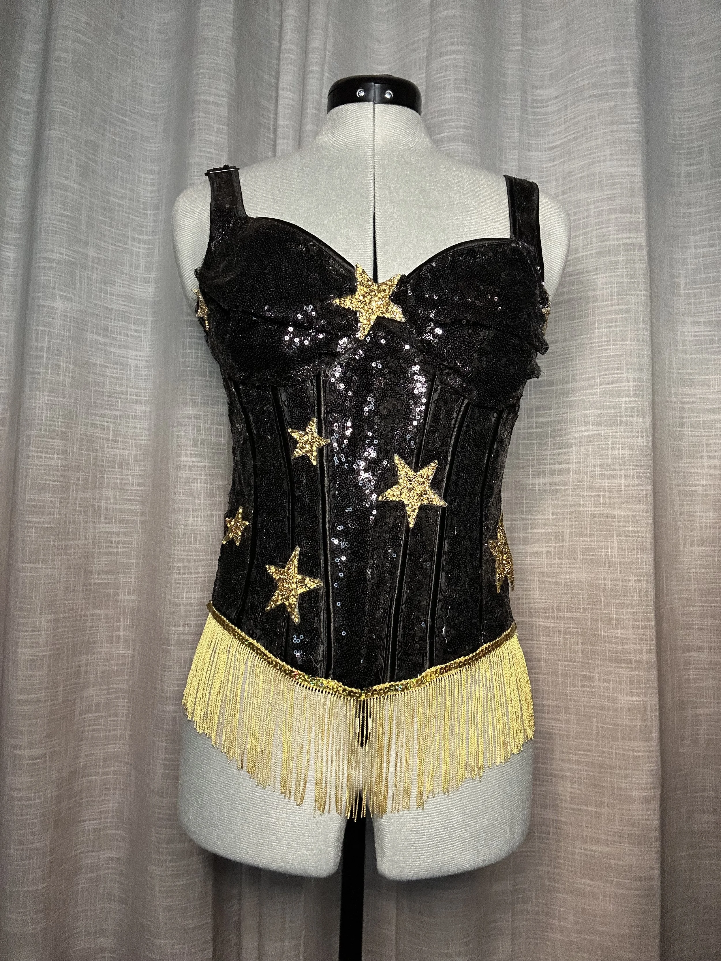 A mannequin dressed in a black and gold star-themed costume with gold fringe at the bottom, set against a light-colored curtain background.