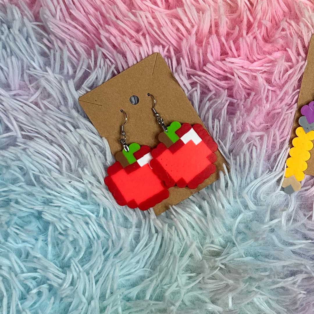 Pixelated strawberry-shaped earrings on a brown card, resting on a multi-colored fluffy surface.