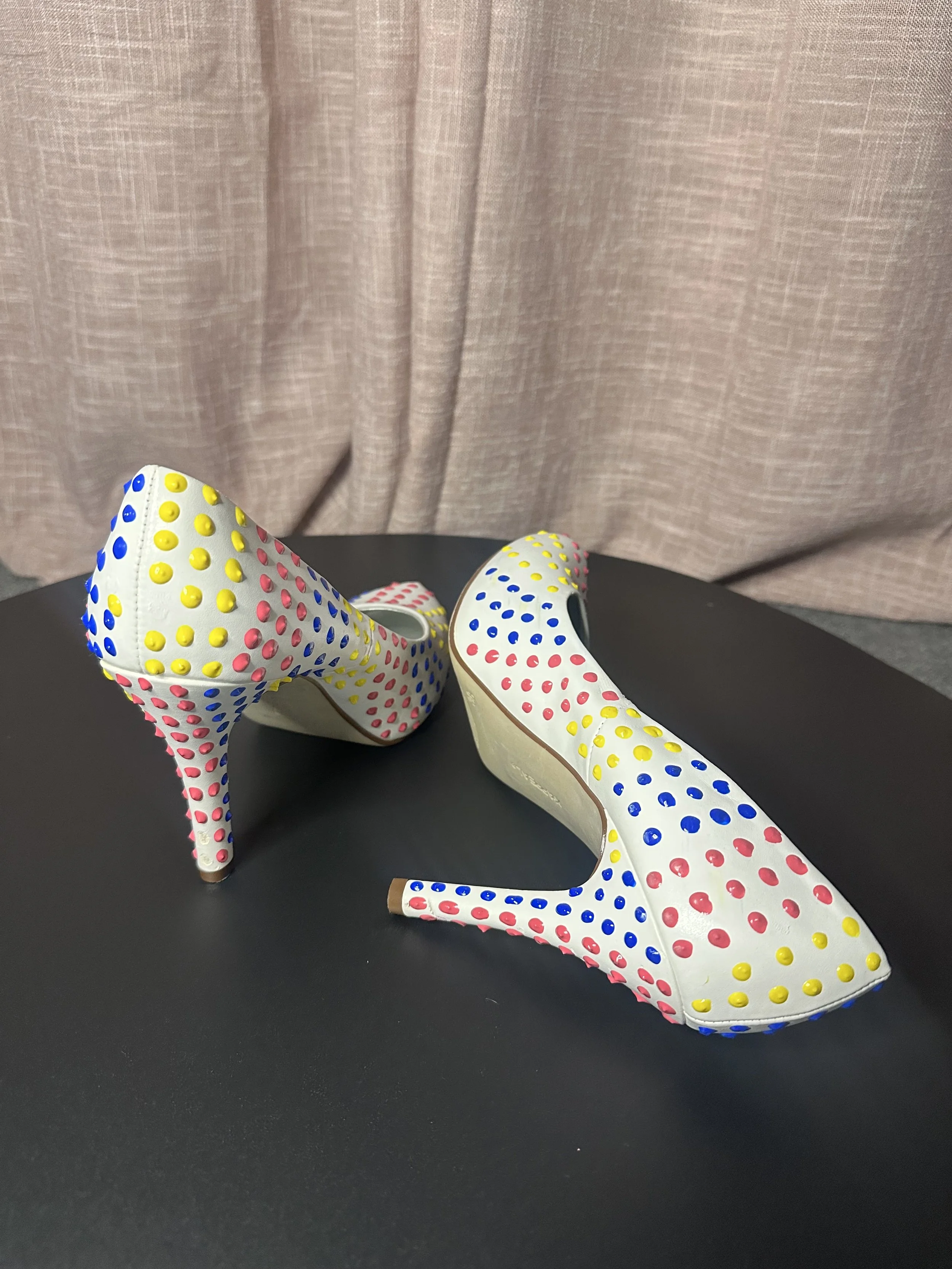 A pair of high-heeled shoes with a white base and colorful polka dots in yellow, pink, and blue, positioned on a black surface with a plain pinkish background.