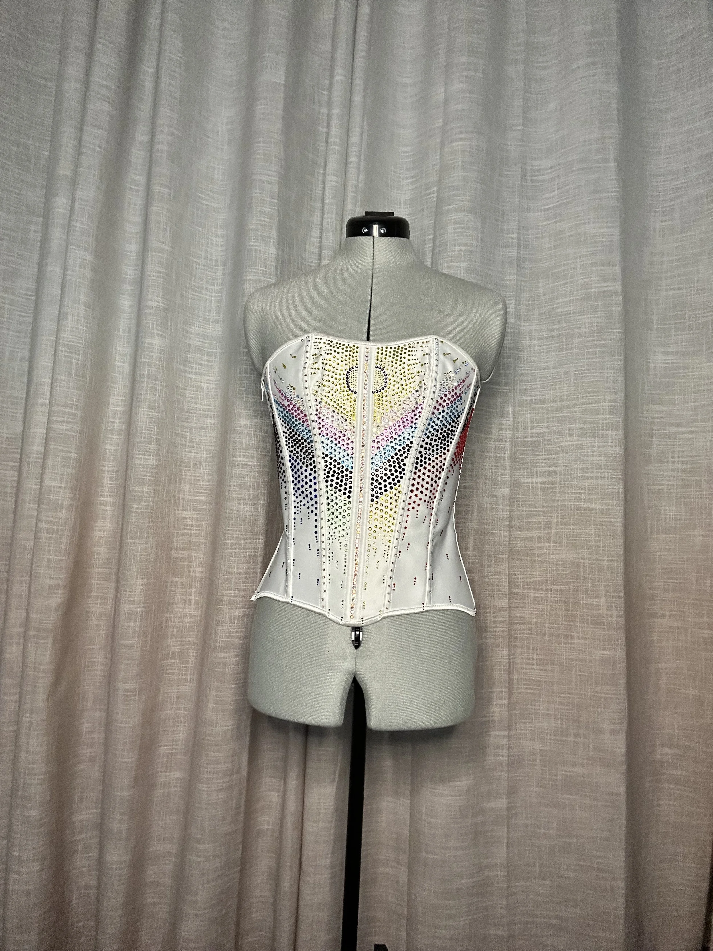 A white corset decorated with multicolored rhinestones arranged in the Progress Flag pattern, displayed on a mannequin.