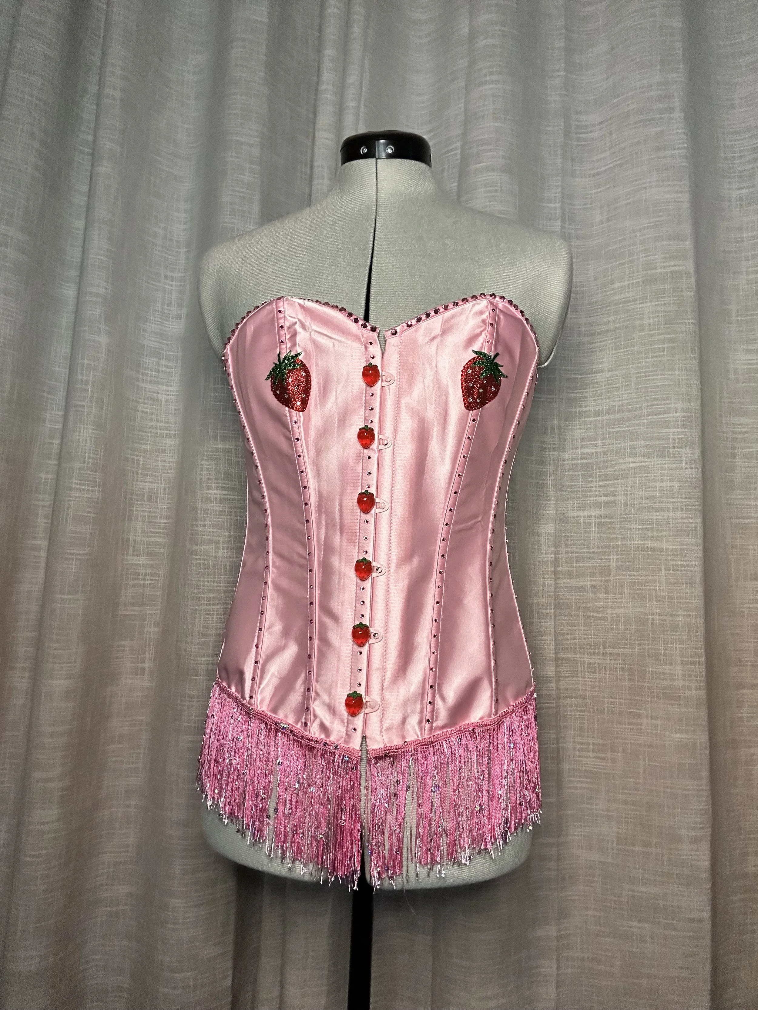 Pink satin bustier with strawberry and strawberry-shaped embellishments and fringe at the bottom, displayed on a mannequin against a curtain background.