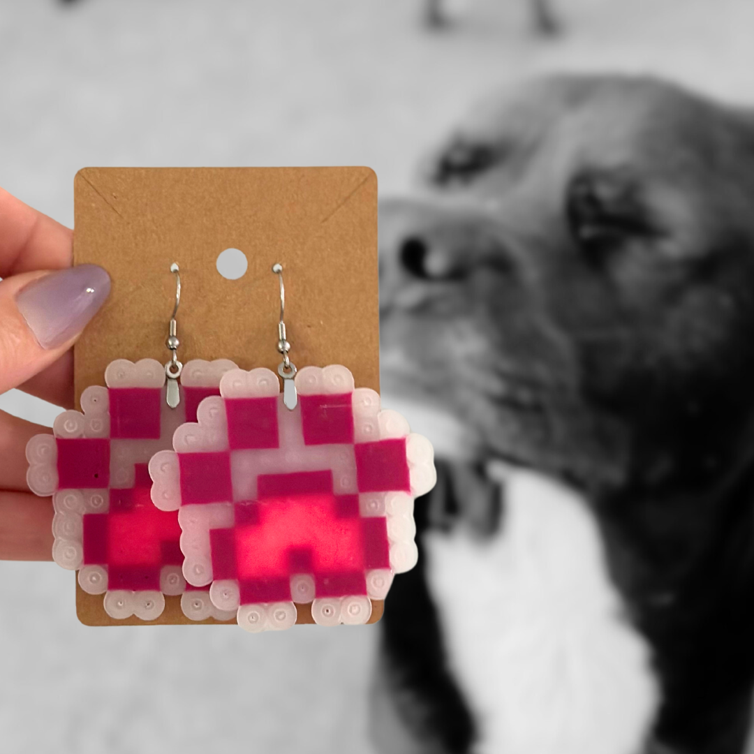 A person holding a cardboard display of pixel art paw print earrings, with a dog blurry in the background.
