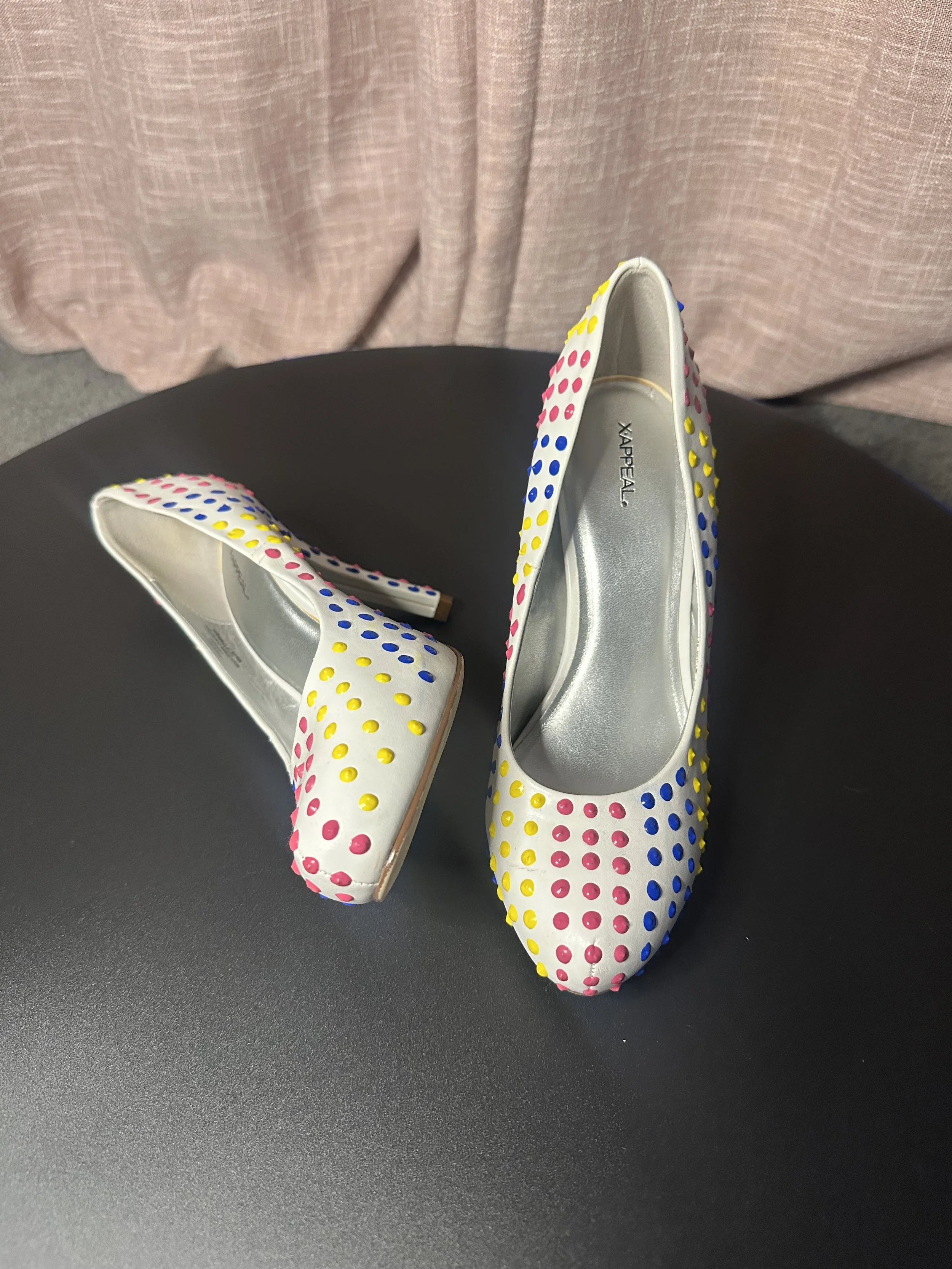 A pair of white high-heeled shoes with colorful polka dots on a black surface.