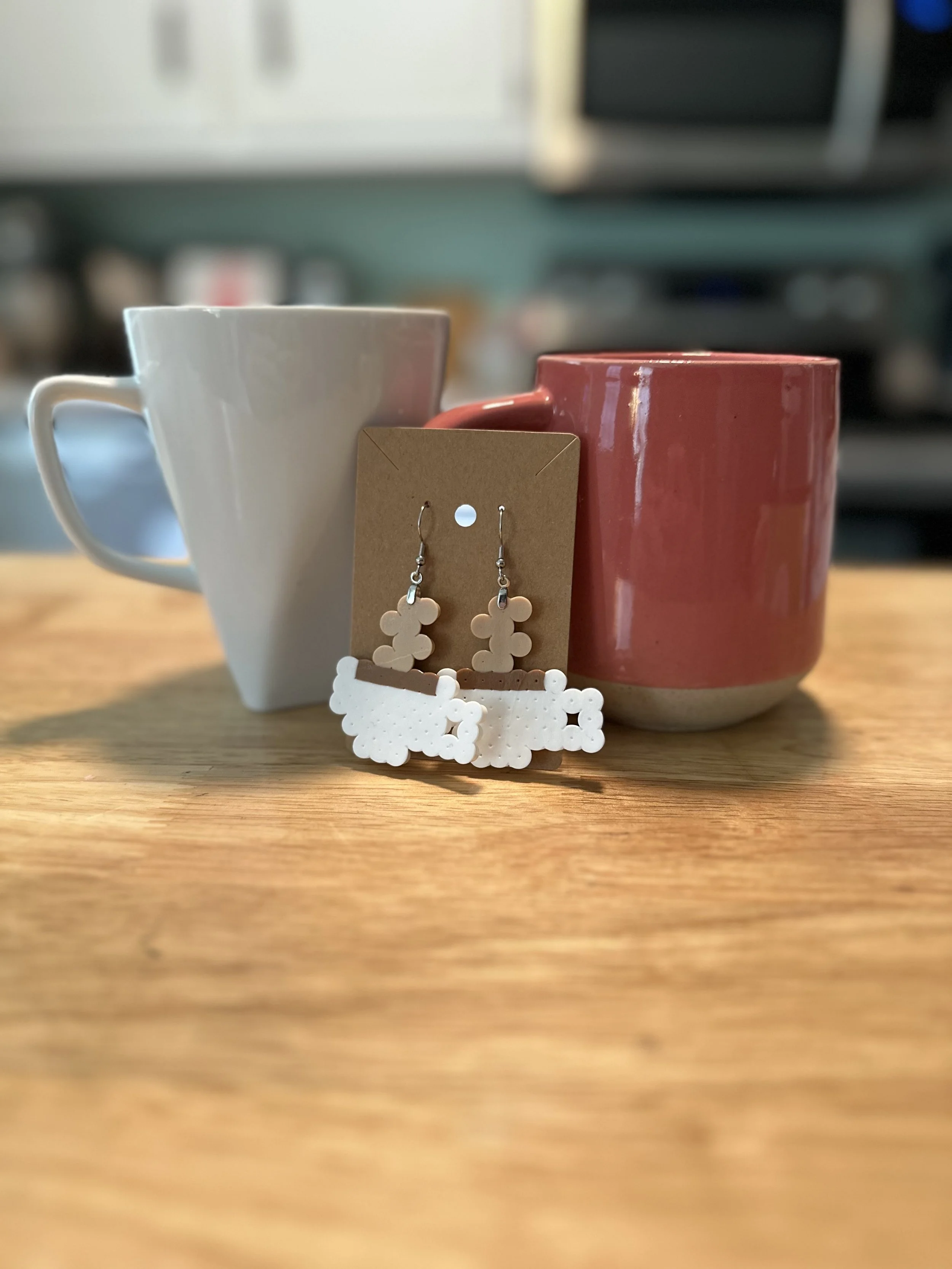 A pair of white earrings with coffee cup-shaped designs hanging on a tan display card, placed between two coffee mugs on a wooden surface.