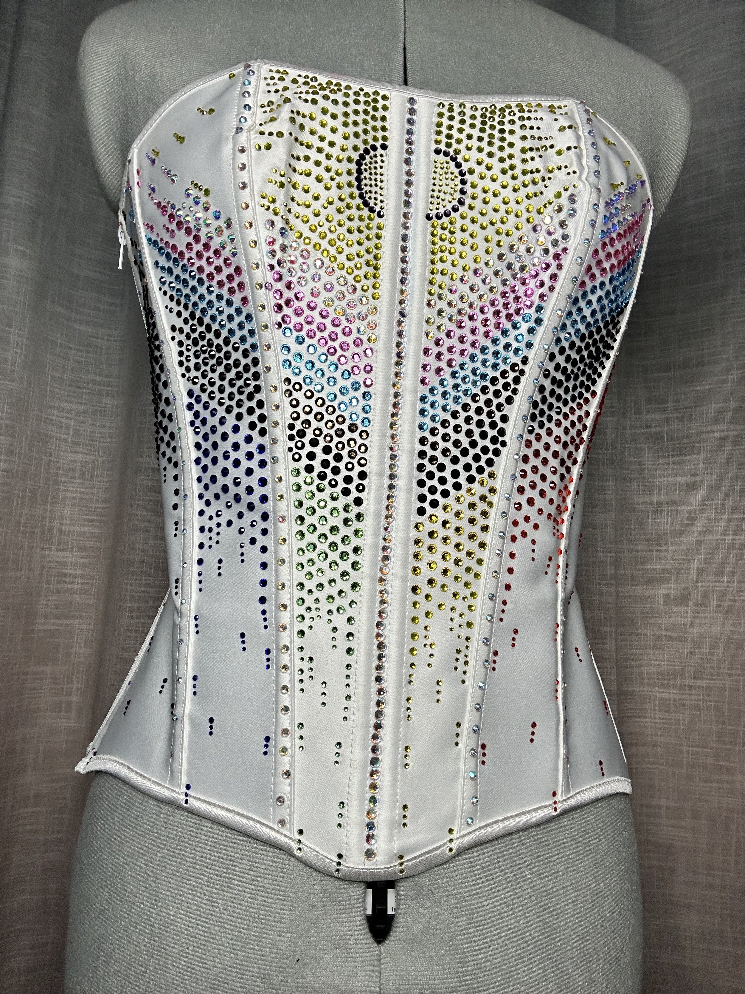 A white corset decorated with multicolored rhinestones arranged in the Progress Flag pattern, displayed on a mannequin.