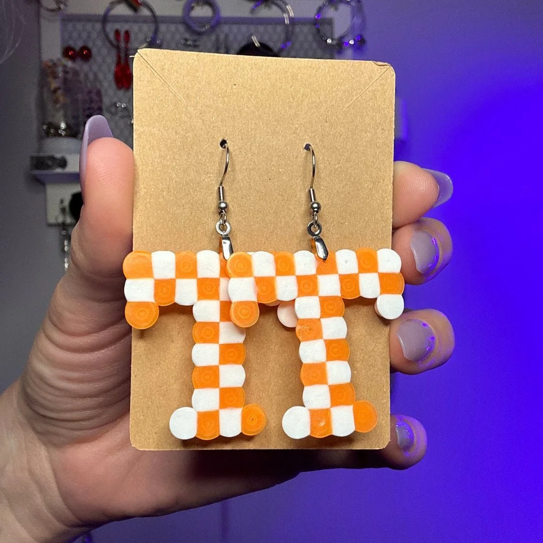 Orange and white checkered earrings in the University of Tennessee shape on a brown card, held by a hand with painted nails.
