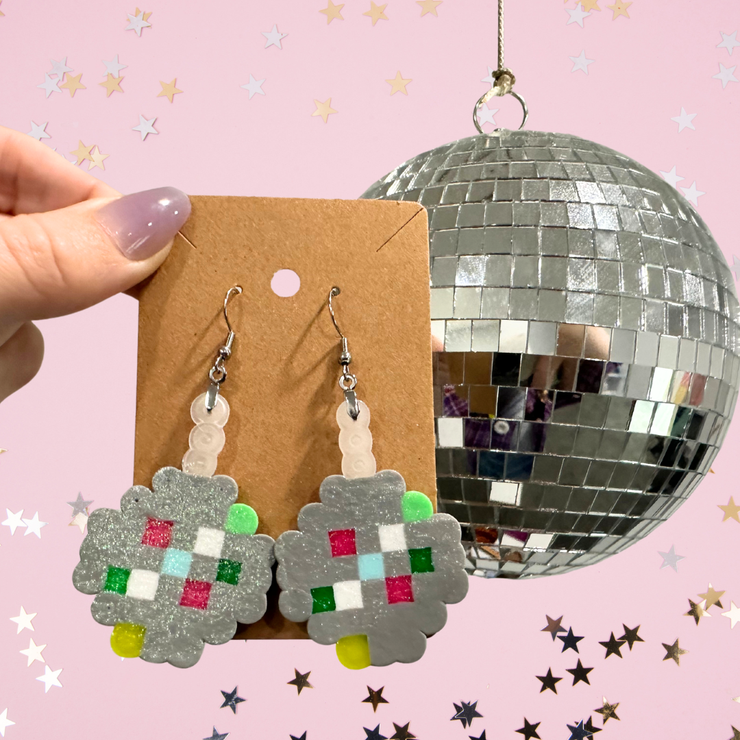 Star-shaped earrings resembling pixelated art, hanging on a brown card, with a disco ball in the background against a pink backdrop decorated with star-shaped confetti.