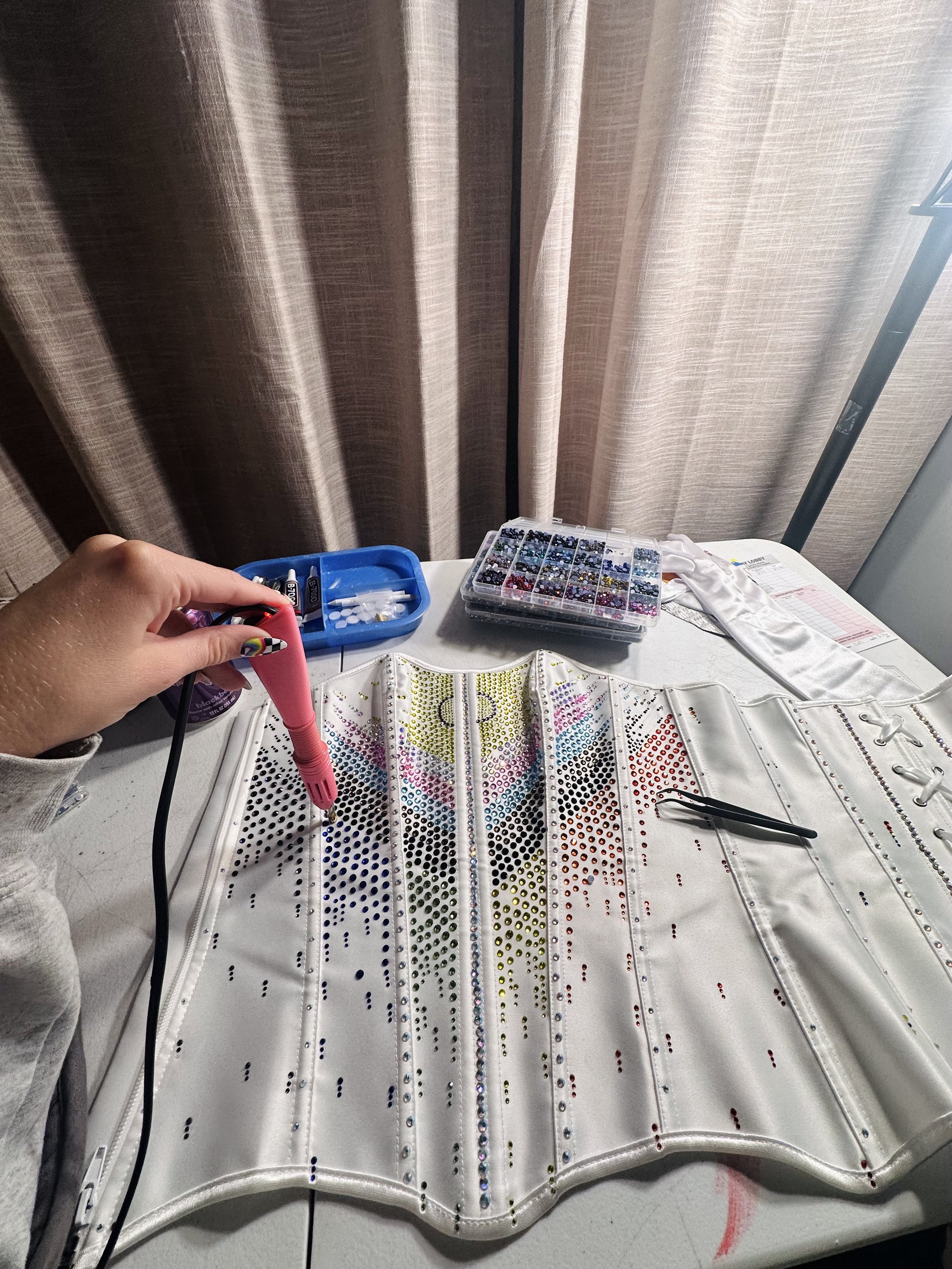A person's hand is holding a heat tool near a rhinestone design on a white fabric surface, with containers of colorful rhinestones and a paper with a chart on a table. The backdrop is a beige curtain.