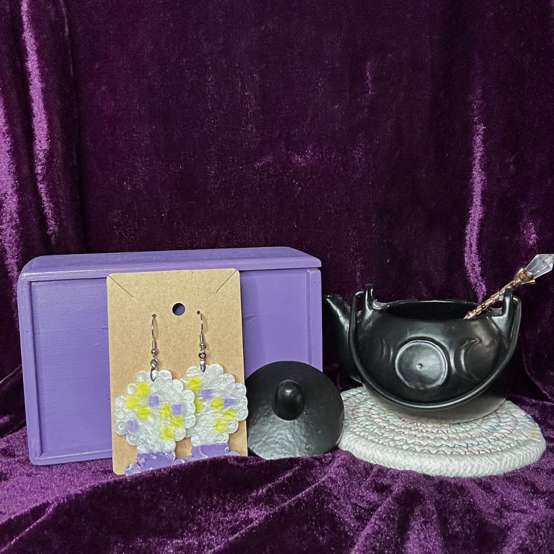 A small jewelry box with purple exterior, a pair of beaded flower earrings on a beige card, a black round object, a witch's cauldron with a wand inside, all placed on a purple velvet background.