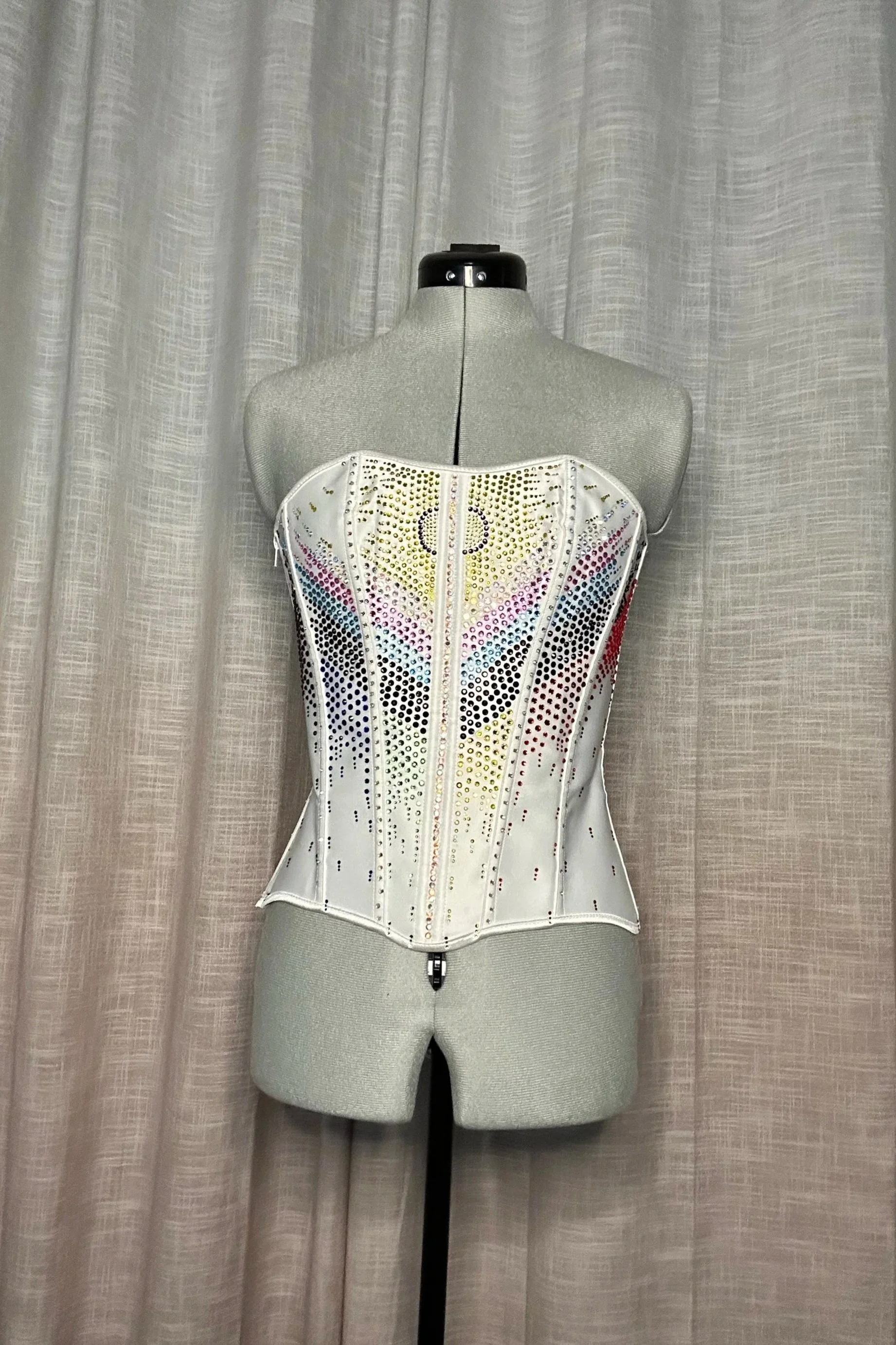 A mannequin wearing a multicolored rhinestone corset with a beige textured curtain background.